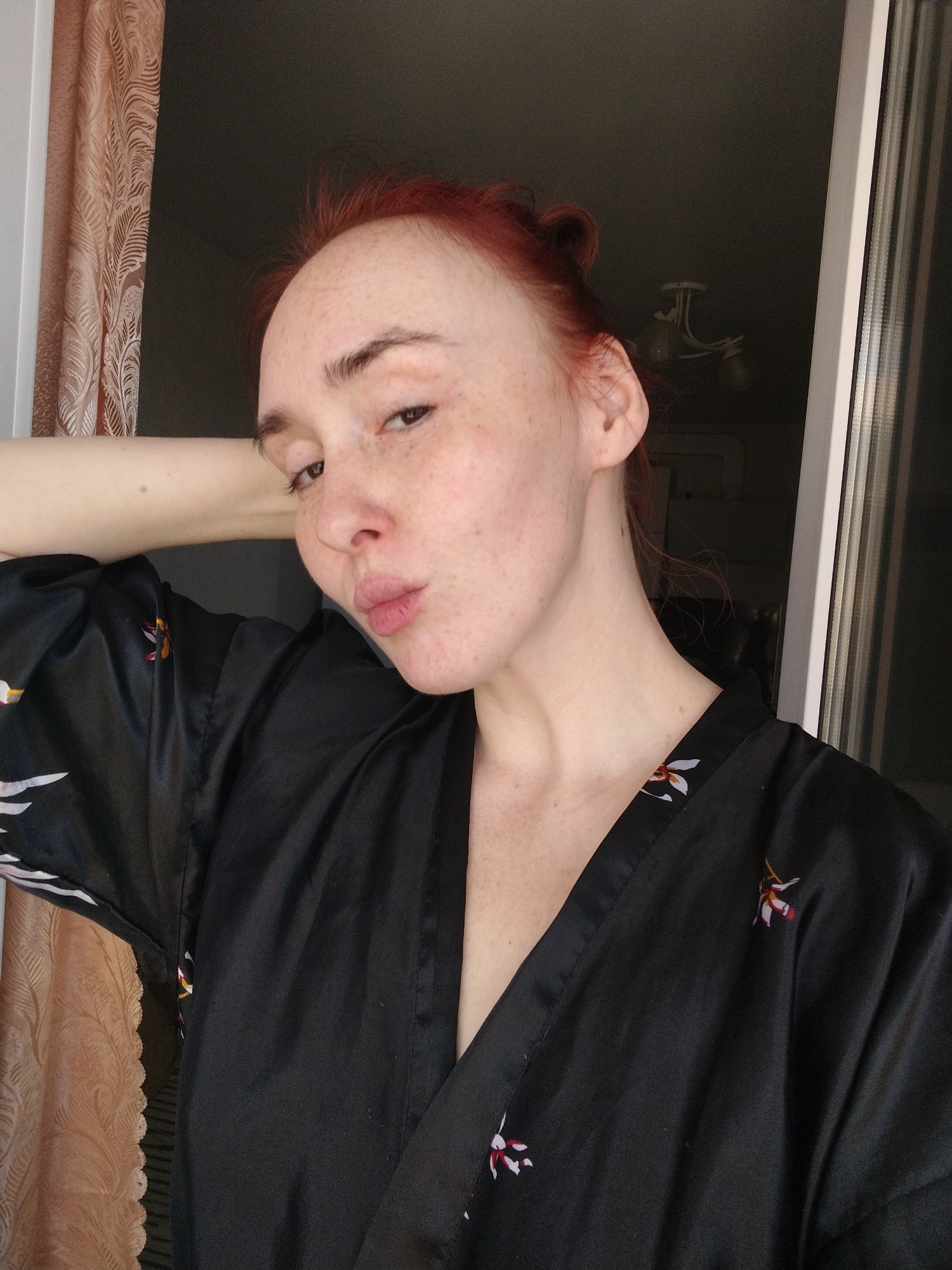 A selfie shows a fair-skinned person with red hair pulled back into a bun, wearing a black robe with floral patterns. The person is looking at the camera with pursed lips. They are positioned in front of a window, with a peach-colored curtain visible on the left. A glimpse of a light fixture can be seen through the window. There is no direct indication of the location being in Izhevsk, Russia.