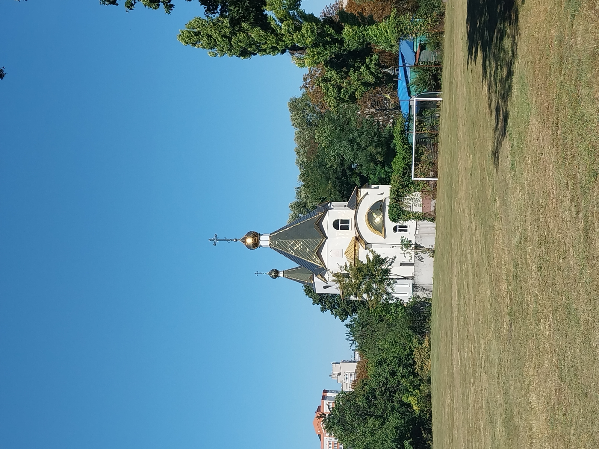 In the city of Doslidne, Ukraine, a white church with a gold-domed roof and a green roof stands prominently on the right side of the frame. The church features arched windows and crosses atop its spires. In the foreground, a vast, dry, mowed grassy field stretches out, casting shadows from unseen trees. To the left of the church, a line of trees and buildings are visible in the background, and a soccer goal is situated on the right side of the field, behind a blue-roofed structure. The sky above is a clear, bright blue.