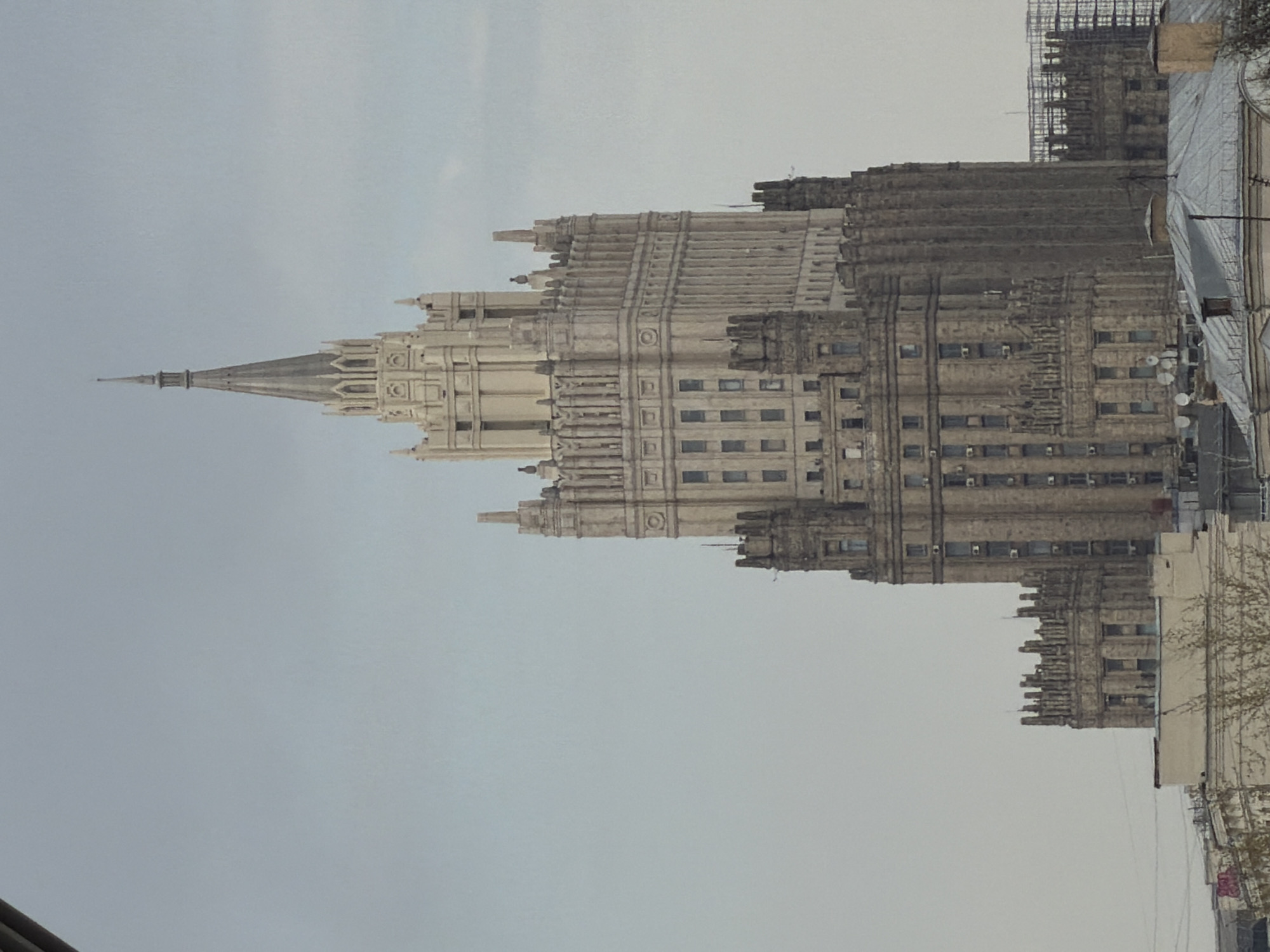 The image shows the Ministry of Foreign Affairs building, one of the Seven Sisters skyscrapers, located in Moscow, Russia. The building is depicted from a slightly elevated position, with a cloudy sky in the background. The architectural details of the skyscraper are visible, including its tiered structure, spires, and windows. Scaffolding is partially visible on another building nearby. Other adjacent buildings and some power lines are visible.