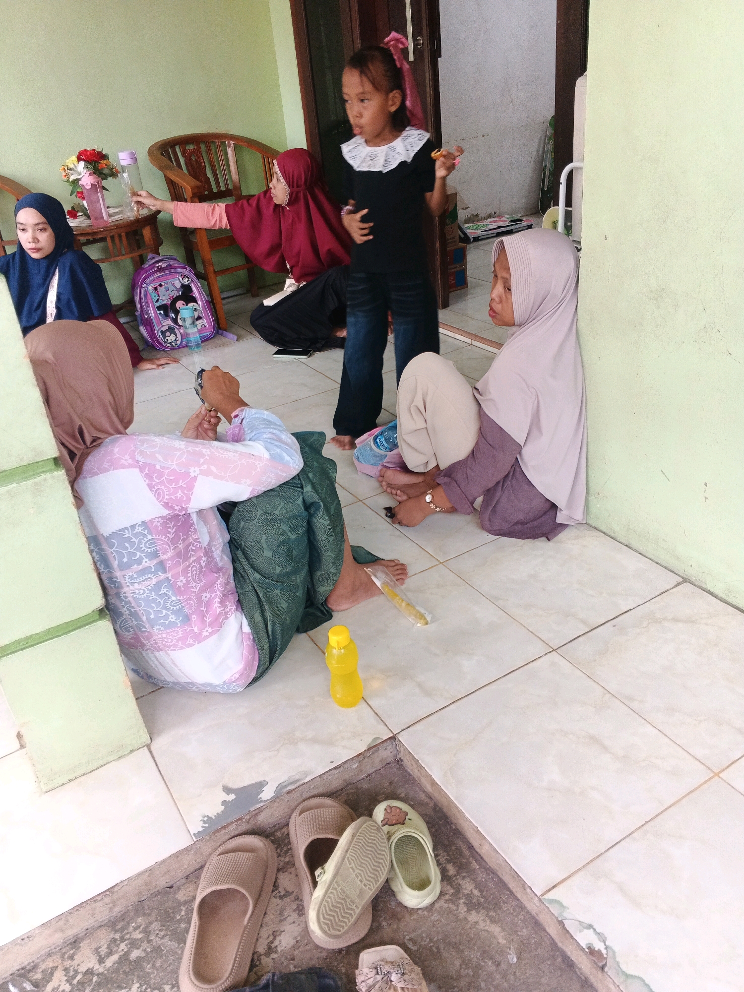 The image depicts a scene at an outdoor dwelling in Kadu, Indonesia, likely a home or community space. The setting is characterized by a tiled floor and a light green wall. In the foreground, several pairs of sandals and slippers are placed on a concrete ledge.
There are at least five individuals visible in the image, all appearing to be female and of varying ages. One young girl, dressed in a black top with a white lace collar and blue jeans, stands near a doorway, holding a small object in her hand. To her left, a woman wearing a maroon hijab and a long-sleeved shirt is seated on the floor, reaching towards a table. Another woman, in a light purple hijab and a purple top, is seated against the green wall, her legs drawn up. Further to the left, a woman in a dark blue hijab is also seated on the floor. Partially visible in the lower left is another woman, seated, wearing a brown hijab and a pink and white patterned top.
Objects present include a purple backpack with cartoon characters, water bottles, and a vase with flowers on the table. The environment suggests a casual, informal gathering. The lighting is natural, indicating daytime. The overall impression is one of a domestic or community gathering, possibly during a break or a pause in activities.