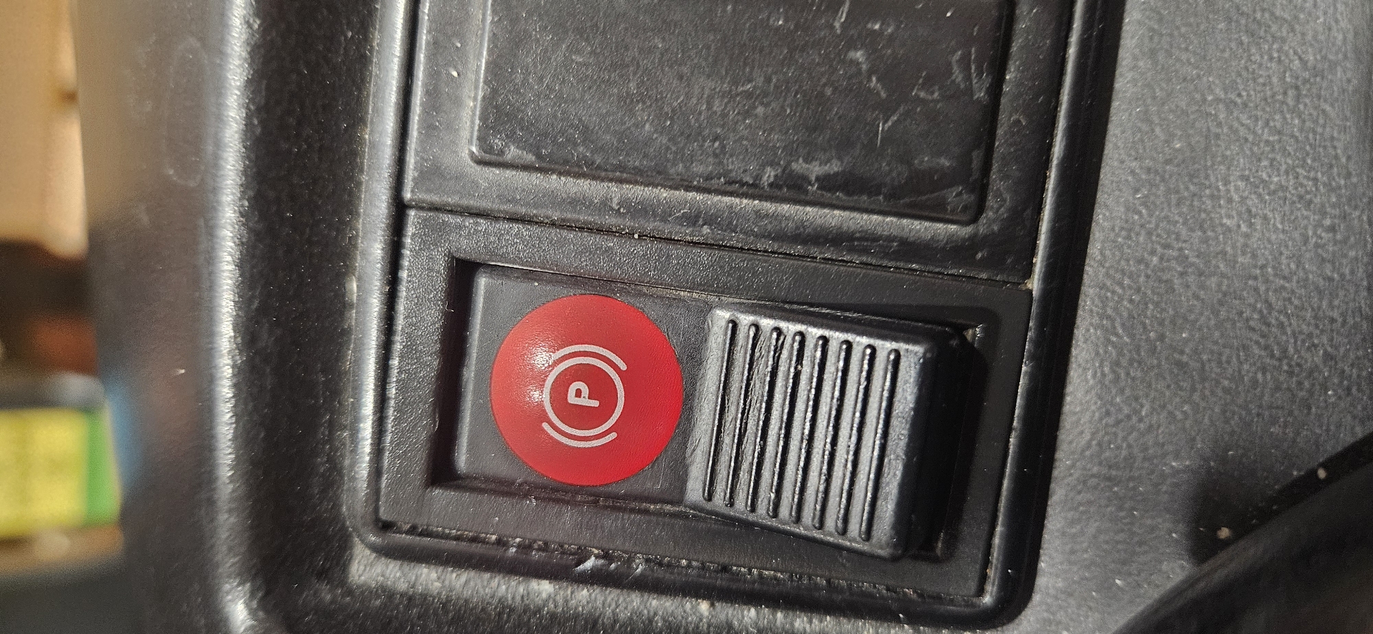 A close-up shot shows a black plastic panel, possibly from the interior of a vehicle. The panel has a rectangular cut-out containing two elements: a red circular button with a white "P" inside two concentric circles (likely indicating a parking brake function), and a black sliding switch with vertical ridges. The panel appears slightly dusty or worn. The location is not determinable from this image.
