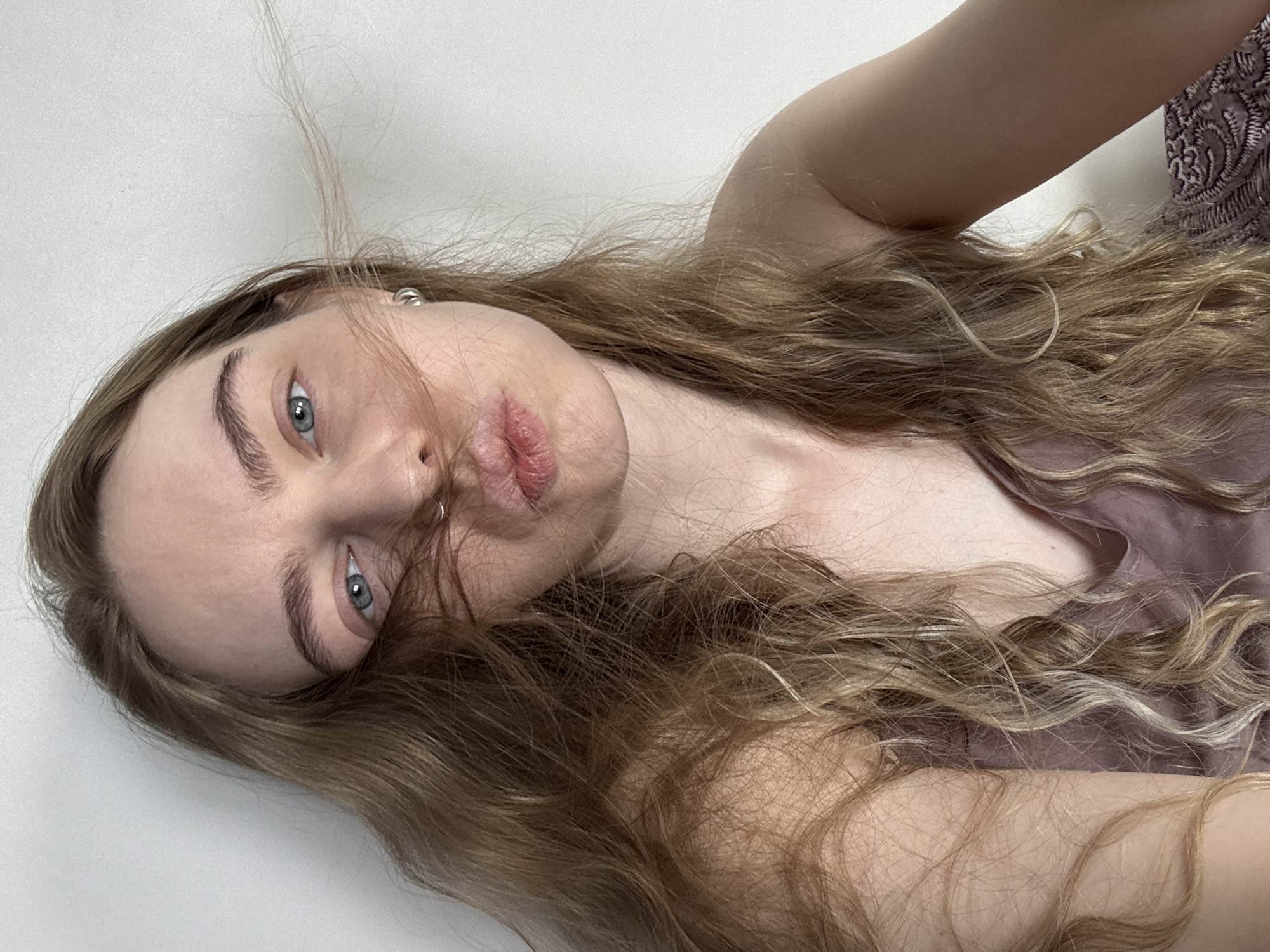 A fair-skinned individual with long, wavy blonde hair and light blue eyes is positioned in front of a white wall. The person is tilting their head to the side. The individual has a piercing in their nose and is wearing what appears to be a light purple top. There is no identifiable location in Krasnoyarsk, Russia.