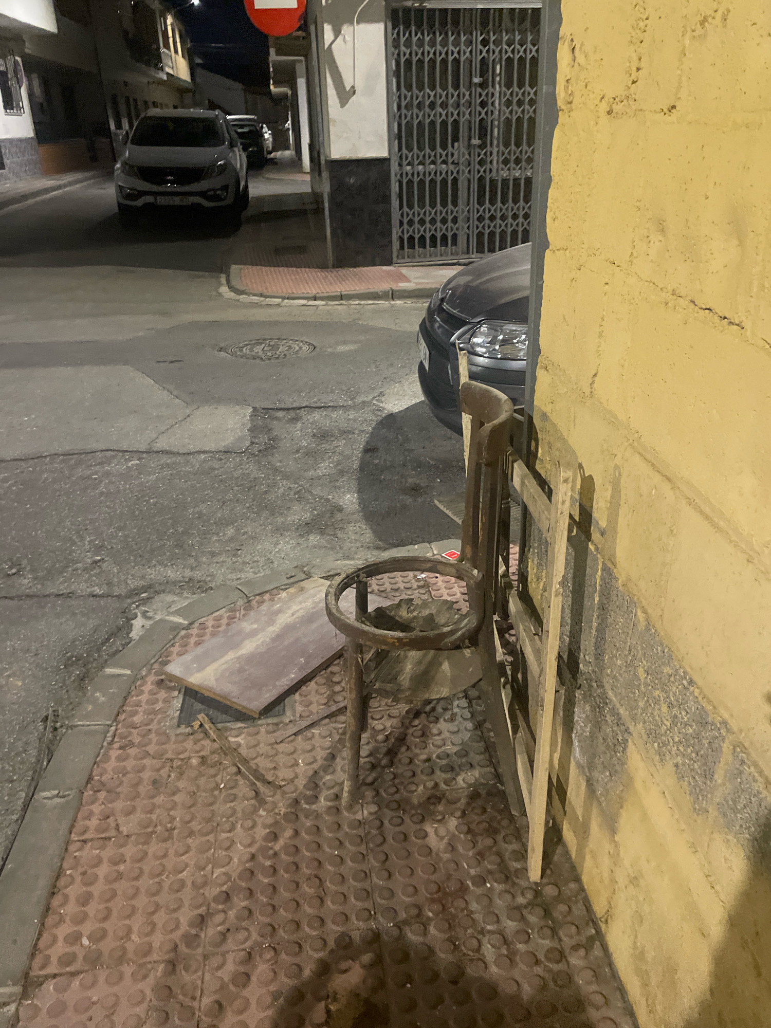 The image depicts a street scene at night in Atarfe, Spain. A white SUV is parked on the left side of the street, with another dark car visible behind it. In the foreground, a rustic wooden chair with a circular seat frame and a wooden backrest is leaning against a textured yellow wall. Several pieces of discarded material, including a flat wooden board and some broken wooden planks, are scattered on the textured red sidewalk next to the chair. A metal grate door is visible in the background, and a circular manhole cover is present on the asphalt road. The lighting suggests it is nighttime, with artificial light illuminating the scene.