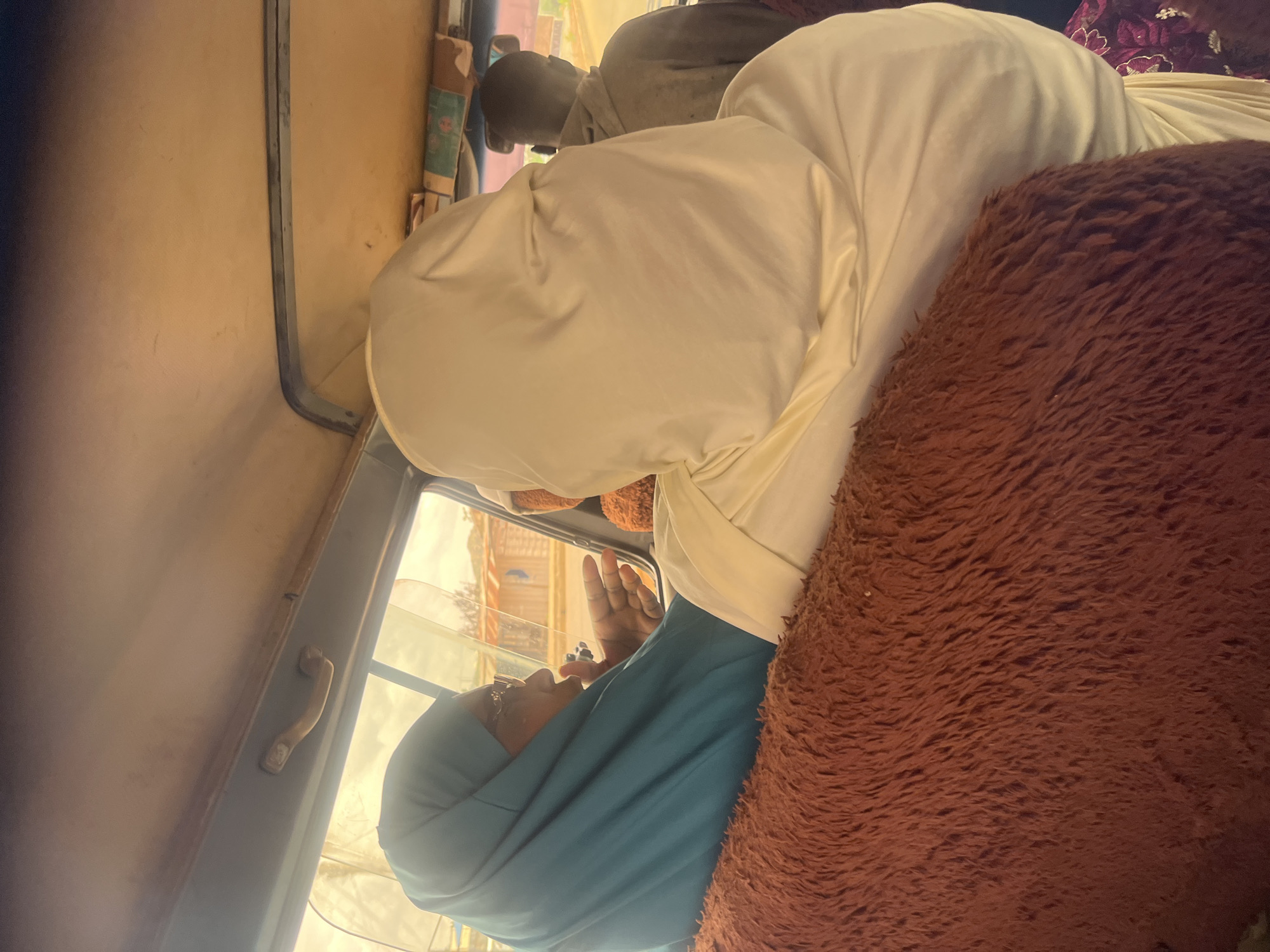 In Bukuru, Nigeria, three individuals are seated inside a vehicle. The foreground is dominated by a shaggy, rust-colored cushion, partially obscuring a person in a cream-colored hijab. To the left, a person wearing a teal hijab and glasses is visible in profile, with their left hand raised. Beyond them, a person in a gray hooded jacket is seated in the back. The interior of the vehicle features a beige headliner and a grey handle. Outside the windows, structures that appear to be small buildings with corrugated metal roofs are visible. The overall lighting suggests daytime.