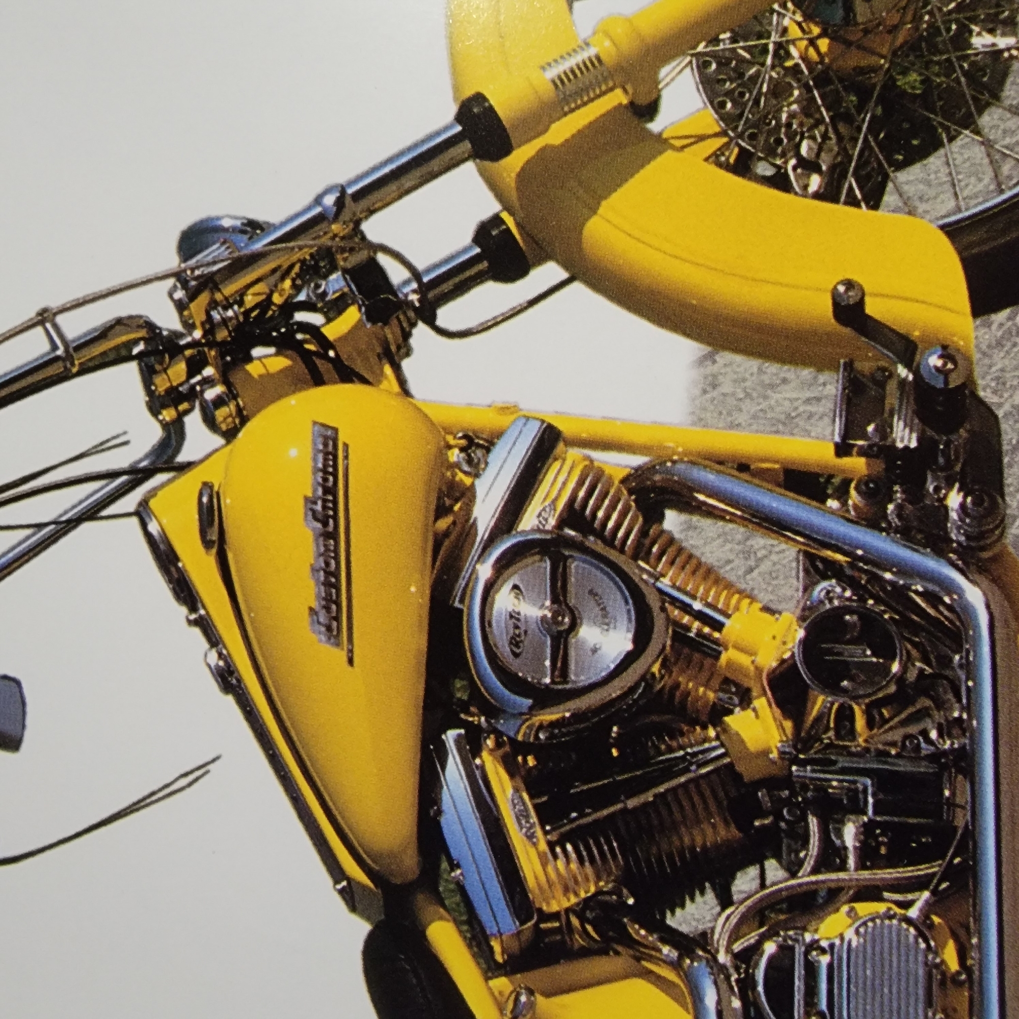 The image shows a close-up, eye-level view of the front and side of a yellow custom motorcycle. The motorcycle has a prominent yellow gas tank with a chrome emblem that reads "Custom Chrome". The engine is visible, with chrome accents and black cylinder fins. The front forks and handlebars are also visible, featuring chrome components. A yellow front fender partially obscures the front wheel, which has chrome spokes and a disc brake. The background is a light, neutral color, suggesting an outdoor setting with soft lighting. There are no people visible in the image, and the focus is solely on the motorcycle itself. The scene appears to be a studio shot or a well-maintained display.