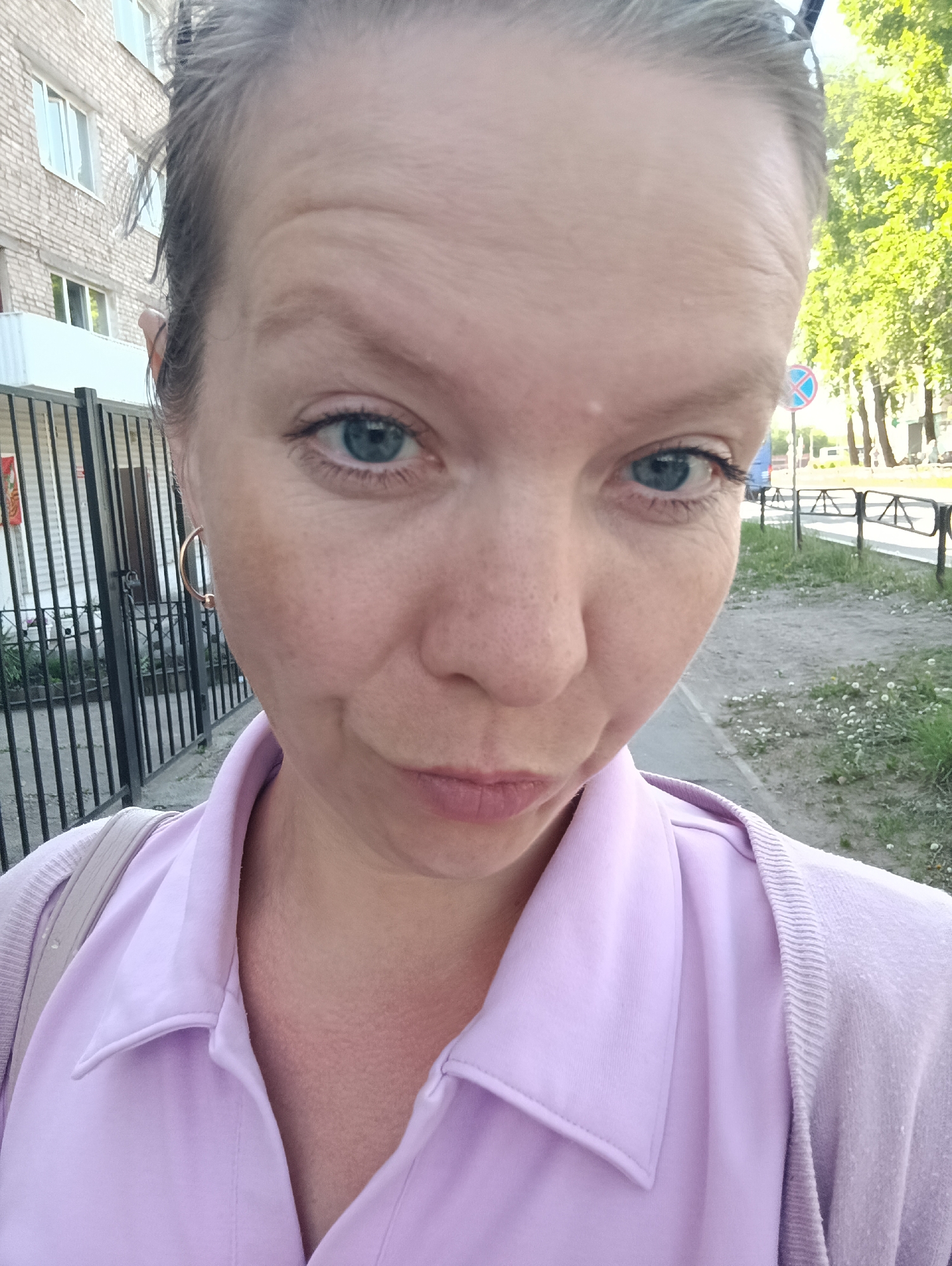 The image is a selfie of a middle-aged woman, outdoors. She is fair-skinned, with blue eyes, and her hair is pulled back. She is wearing a light purple button-down shirt and a matching cardigan. The setting appears to be a city street in Chaikovsky, Russia, as the background shows a building with a brick facade, a black metal fence, a sidewalk, and trees. A road sign is visible in the distance.