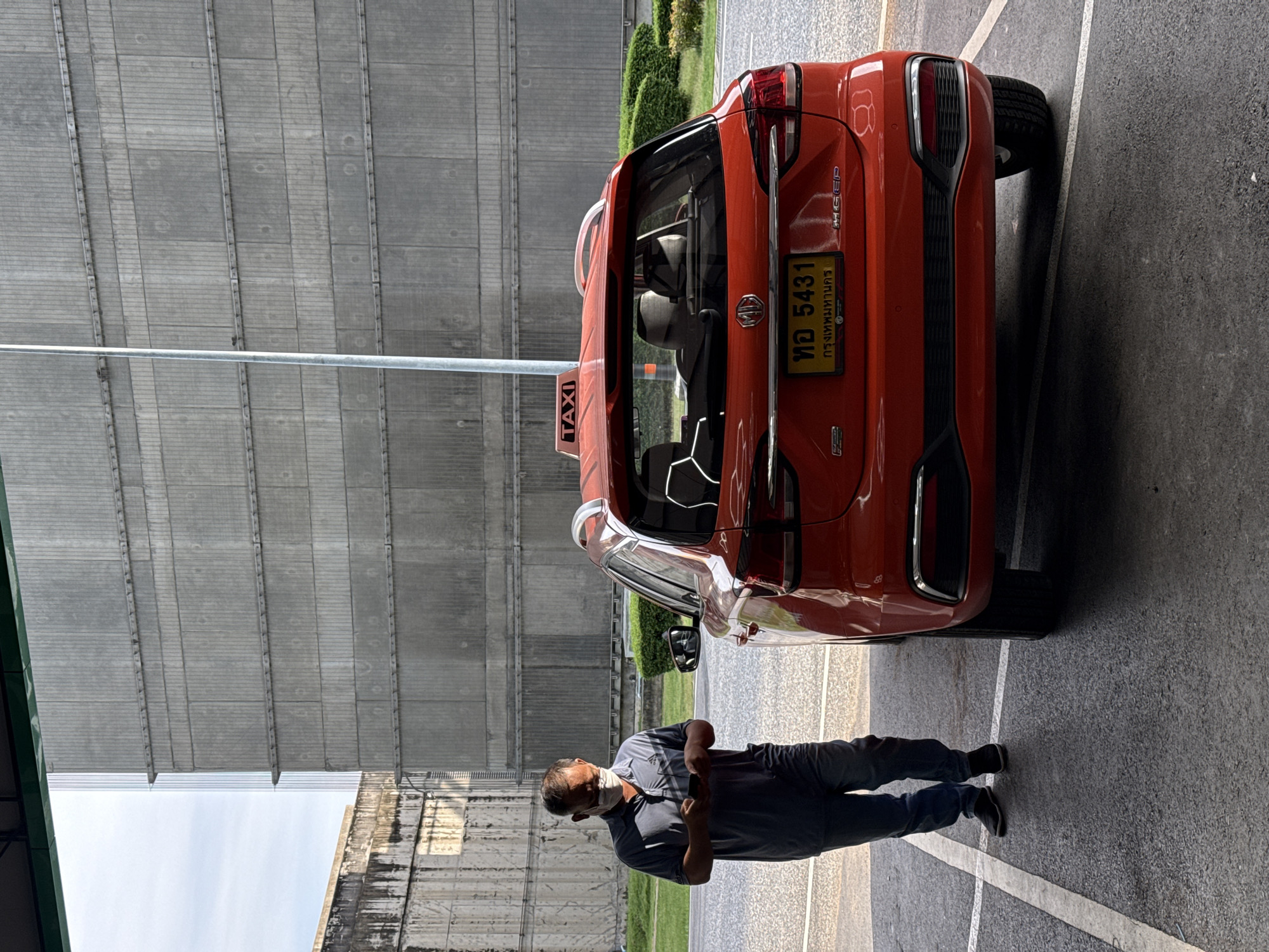 A man wearing a face mask, dark blue t-shirt, and jeans stands beside a red MG electric taxi. The taxi has a "TAXI" sign on its roof and a yellow license plate with Thai characters and the number "5431". The man is looking at his phone. In the background, a large, grey, industrial building with a textured facade and horizontal banding dominates the scene, with a smaller, lighter colored building visible to the left. Green bushes are visible in the mid-ground. The ground is asphalt with white parking lines. The scene appears to be in an outdoor parking area, possibly at a transportation hub or industrial complex in Nong Prue Subdistrict, Thailand.