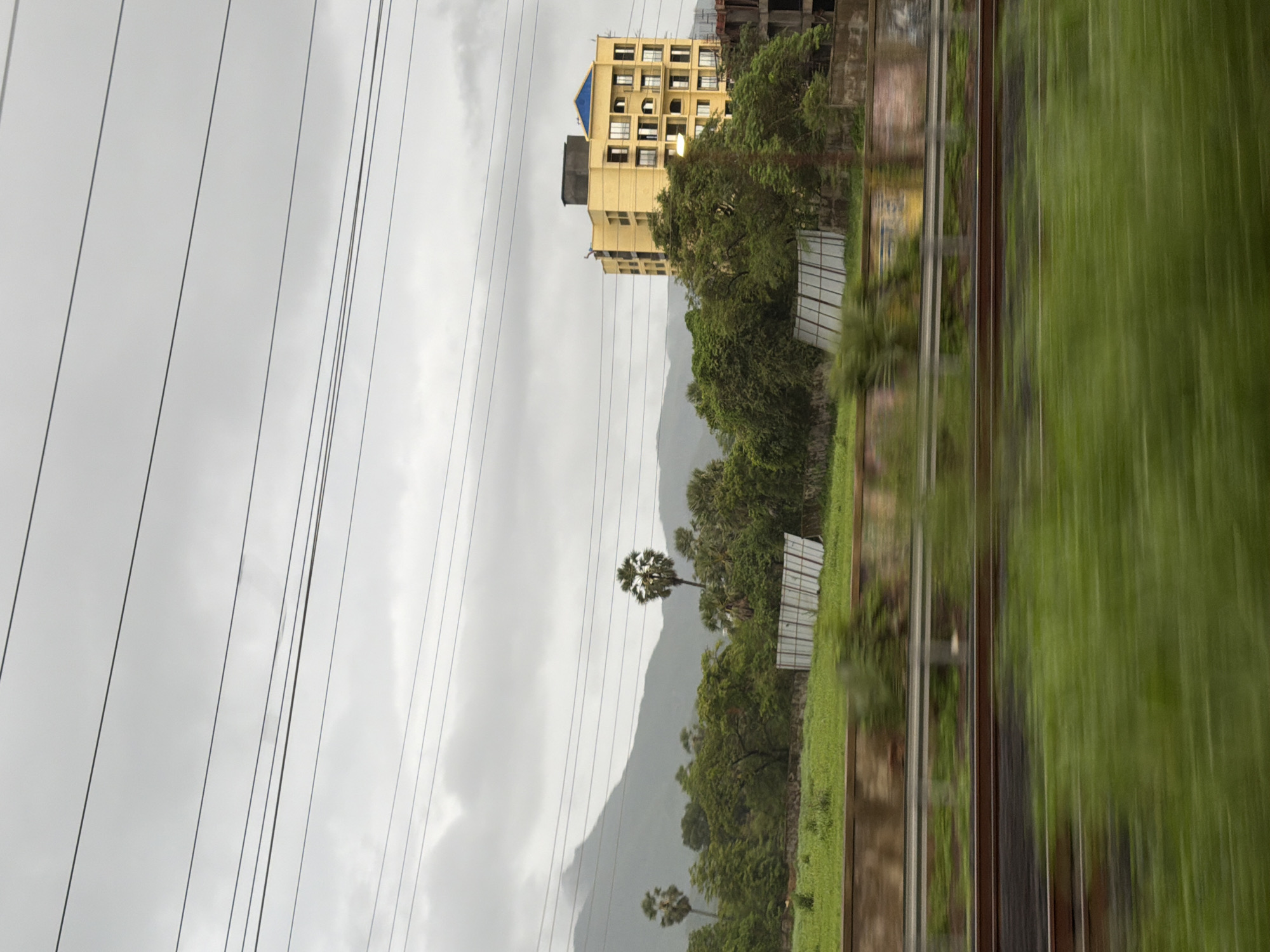 This image captures a view from a moving vehicle, likely a train, in Palghar, India, on a cloudy day. The foreground is blurred by motion, showing green vegetation and train tracks. In the mid-ground, a vibrant yellow, multi-story apartment building with balconies and a blue-tiled roof stands out amidst lush green trees. Behind the trees, a misty mountain range forms the backdrop under a heavily overcast sky. Several power lines crisscross the sky. There are also some white, panel-like structures in the mid-ground, possibly part of a construction or agricultural setup. The overall scene depicts an urban fringe environment with a blend of natural landscapes and developing infrastructure.