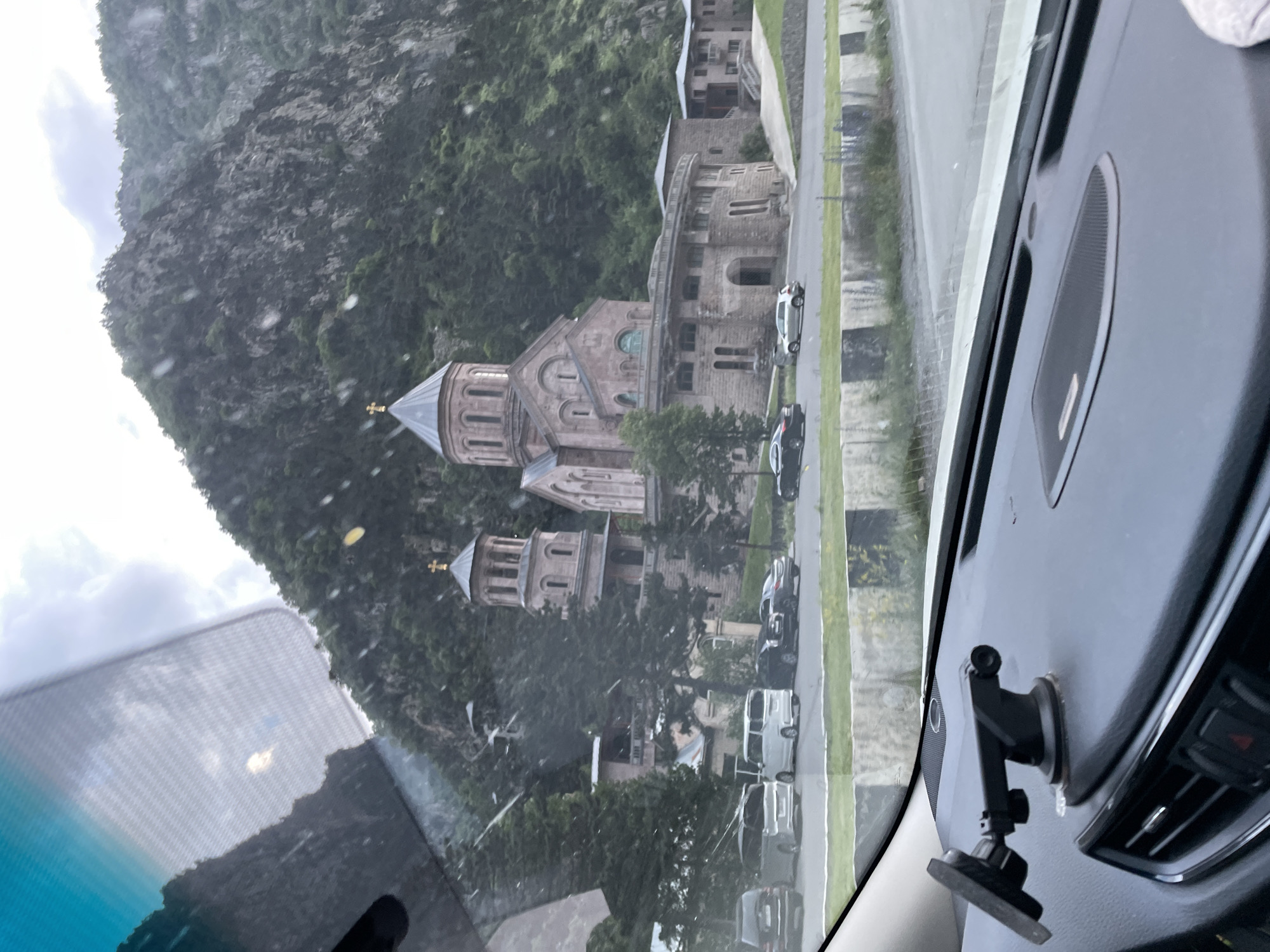 The image, captured from inside a vehicle, shows a view through the windshield of a stone church complex situated at the base of a steep, tree-covered mountain. The church features multiple domes, one prominent central dome topped with a cross, and a tall, separate bell tower also crowned with a cross. The architecture suggests an Orthodox Christian style. In front of the church, several cars are parked on a paved area. The foreground of the image is dominated by the dashboard of the vehicle, including a suction cup phone mount. The environment appears to be overcast with some visible water droplets on the windshield, suggesting rain or a wet surface. The location is identified as Dariali, Georgia, likely near the Dariali Gorge, known for its monasteries and natural beauty.