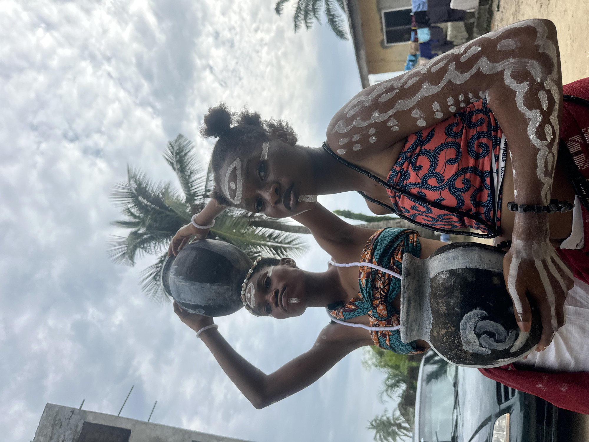 In an outdoor, overcast setting in Badagry, Nigeria, two young women adorned with tribal markings on their faces and bodies are the primary subjects. The woman on the left, with a beaded headband and necklace, balances a large, dark, round pot on her head. She is wearing a brightly colored, patterned fabric wrapped around her torso. The woman on the right, also with tribal markings and dark beaded necklaces, holds a smaller, dark pot with white markings in her hands. She is wearing a strappy, pink and blue patterned top. Both women have white paint applied to their faces, including around their eyes and on their lips. Their bodies are also decorated with white paint. In the background, a partially constructed building is visible on the left, palm trees are scattered throughout, and a car is parked on the left side of the frame. Clothing hangs on a line in the background on the right, suggesting a residential area. The overall scene appears to be a cultural or traditional depiction.
