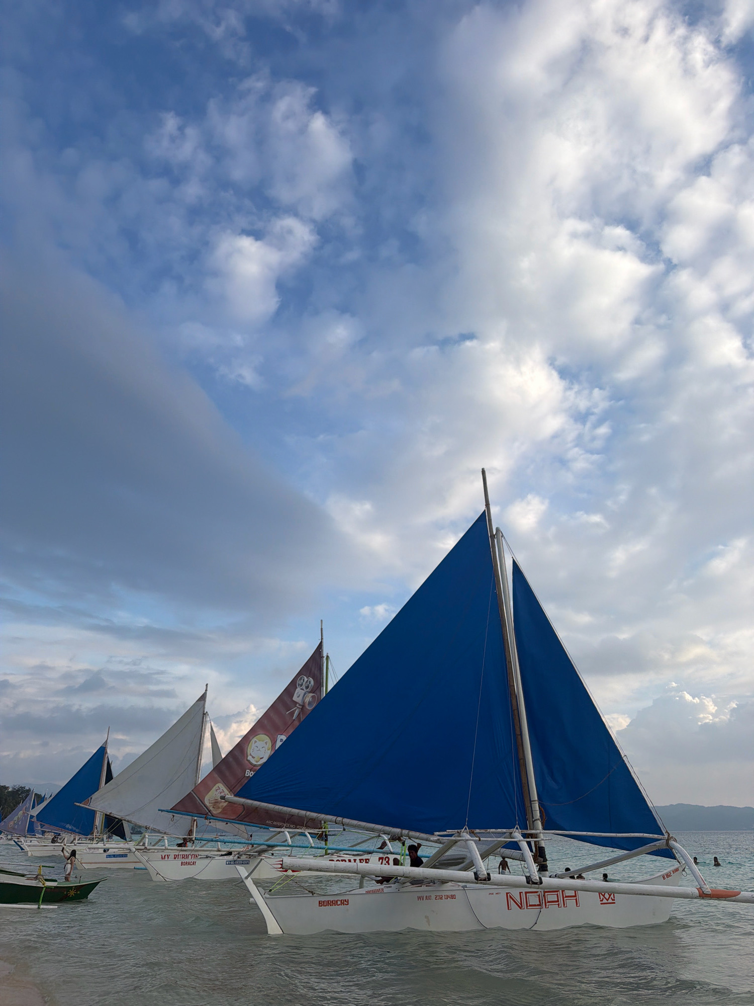 The image depicts a body of water with several sailboats docked or anchored near a sandy shore. The sailboats are traditional outrigger canoes with triangular sails. The nearest sailboat in the foreground has a blue sail and the name "NOAH" written in red on its hull. The sails of other boats are white and brown. The sky is filled with dramatic clouds, with patches of blue sky visible. In the background, landforms and a few people swimming or standing in the water can be seen. The scene suggests a tropical coastal location, likely in the city of Malay, Philippines, given the presence of these types of sailboats, known as "paraw" sailing boats, commonly found in tourist areas like Boracay.