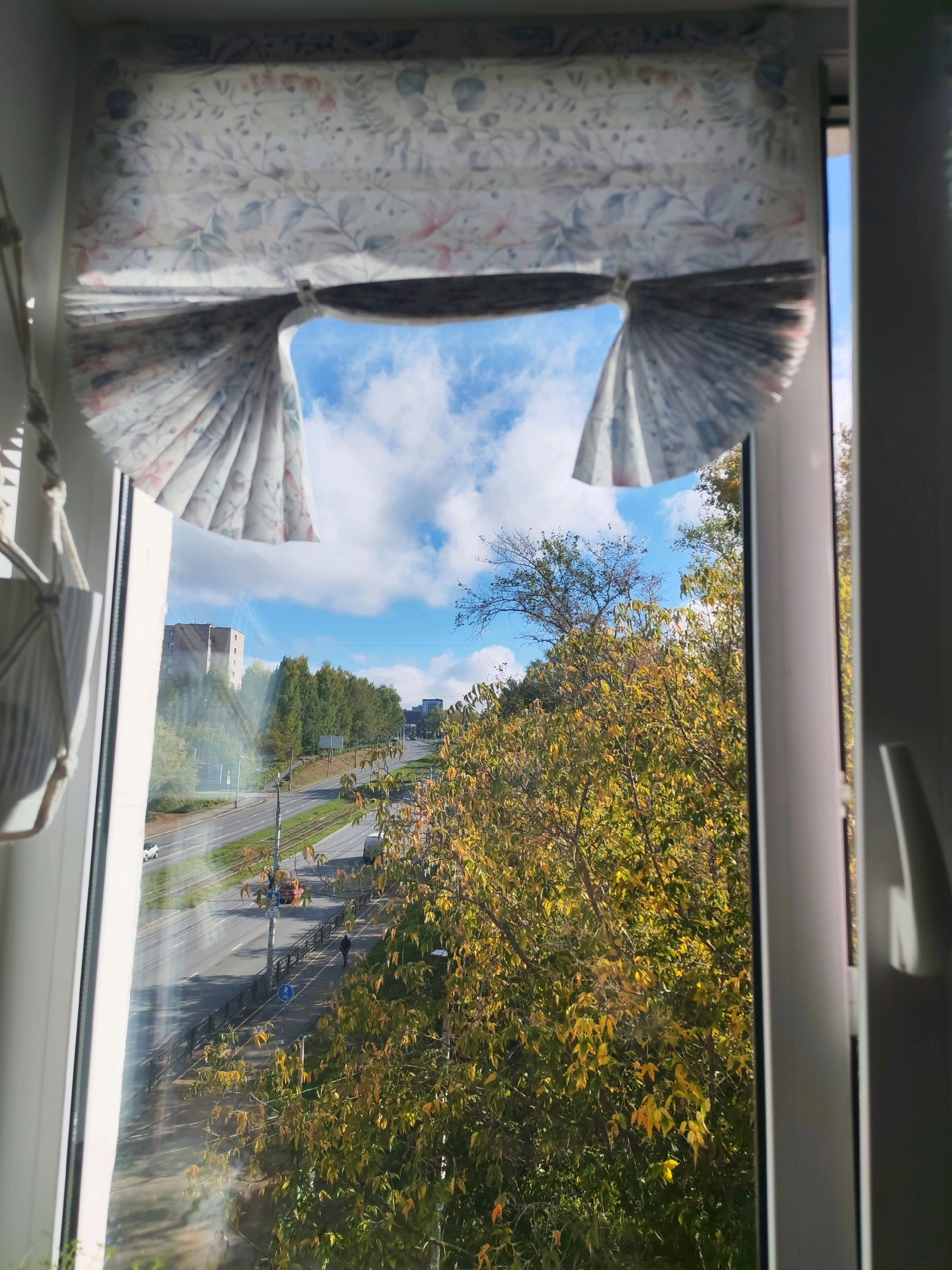 The image shows a view through a window with a decorative roman blind partially drawn. The blind features a floral pattern with muted colors of pink, blue, and green on a white background. The blind is cinched in the middle, creating a pleated effect on the sides.
Through the window, a street scene in Izhevsk, Russia is visible. The time of day appears to be late morning or early afternoon, with clear blue skies dotted with white clouds. Sunlight illuminates the scene, casting shadows.
A multi-lane road runs through the scene, with a tram track visible alongside it. Several vehicles, including cars and a small red truck, are present on the road. A pedestrian walkway runs parallel to the road, bordered by a dark railing. A few pedestrians are visible on the walkway, one of whom is walking a dog.
The surrounding environment includes a hillside covered with trees displaying the colors of autumn, with a mix of green and yellow foliage. A tall, light-colored building is visible in the distance, likely an apartment complex. The overall impression is a view of an urban environment in autumn, seen from a residential window.