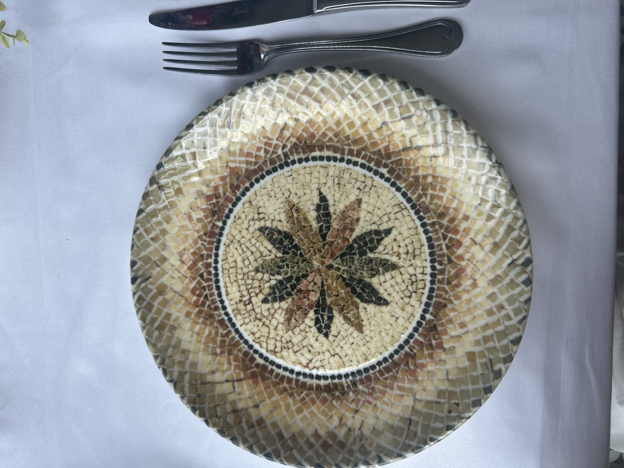 The image shows a place setting on a white tablecloth. A mosaic-patterned plate occupies the center. A fork and knife are placed horizontally above the plate. The scene appears to be indoors. No specific information indicates a location in Başakşehir, Turkey. No people are visible.