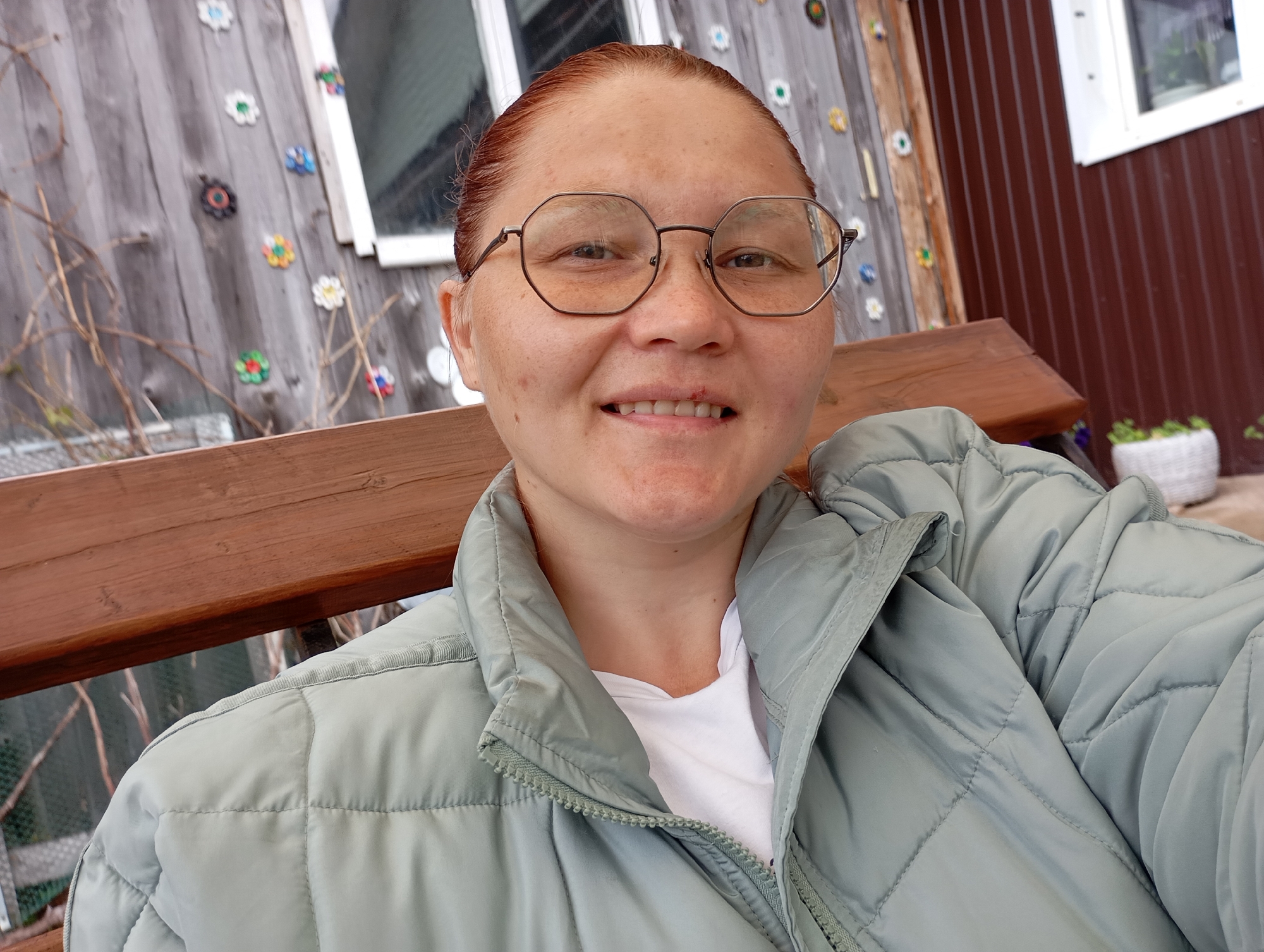 A close-up selfie taken in Фоки, Russia, features an adult female subject seated outdoors. The subject has reddish-brown hair, pulled back, and visible freckles across her face. She is wearing dark-framed, hexagonal eyeglasses and is smiling, revealing her upper teeth. A small reddish mark is present above her upper lip, slightly right of the philtrum. She is dressed in a white t-shirt, partially visible under an open, light sage green quilted puffer jacket with a stand-up collar and a front zipper. She is positioned on a dark brown wooden bench, with a portion of its backrest and armrest visible. The background consists of two distinct building wall sections: to the left, a greyish-brown wooden plank wall adorned with multiple colorful, flower-shaped decorations, with bare branches partially visible in front of it; to the right, a dark brown, vertically paneled wall. Both wall sections feature white-framed windows. A white woven basket containing green plants with purple flowers is visible on the ground near the base of the dark brown wall.