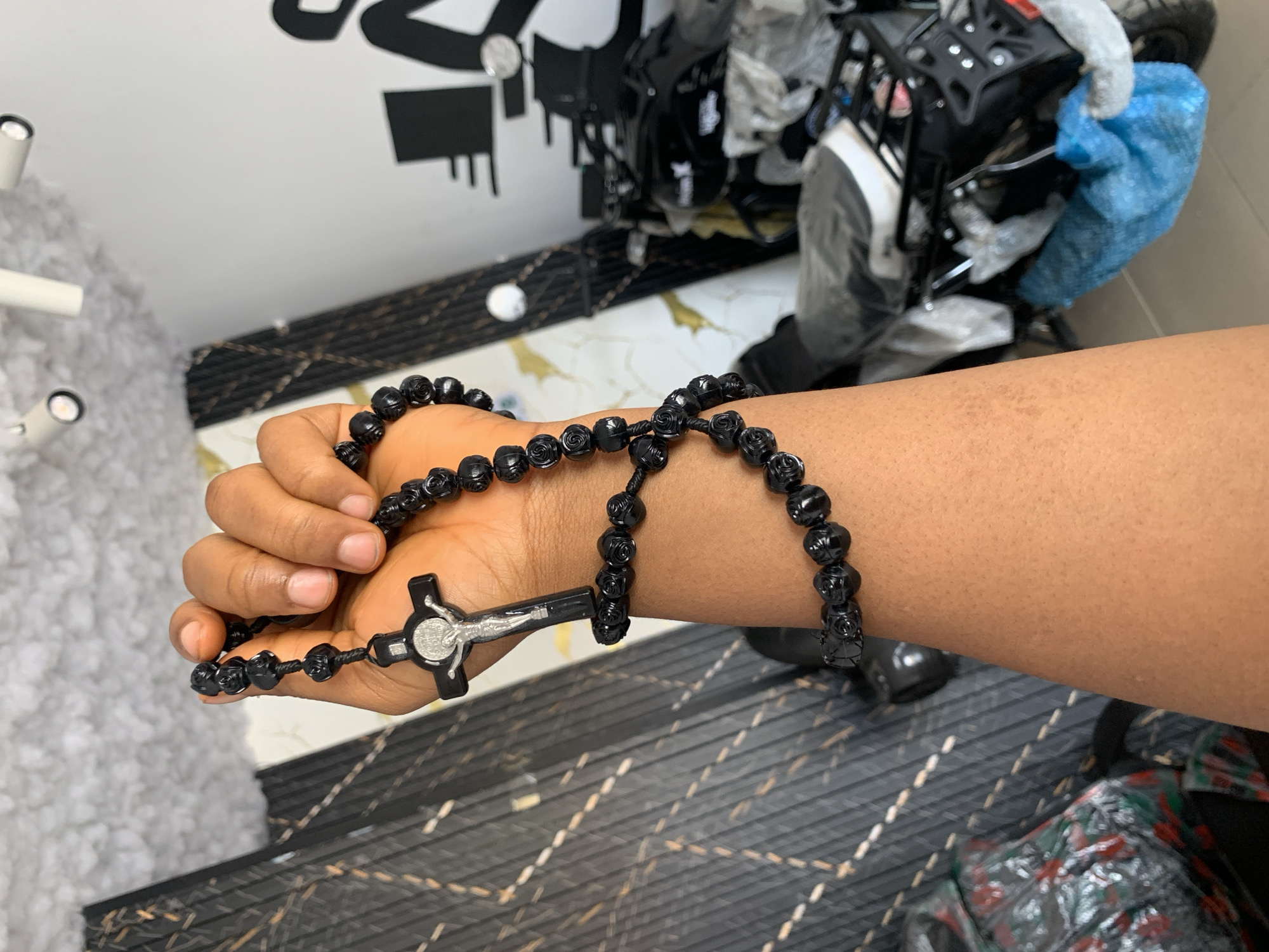 A person holds a black rosary with a silver crucifix. The beads appear to have a rose design. The setting is indoors, likely in a residential or retail space given the patterned flooring and presence of what seems to be packaged items in the background. There is a motorcycle or motorbike part in a plastic package in the upper right of the image. No specific details confirm the location as Jos, Nigeria.