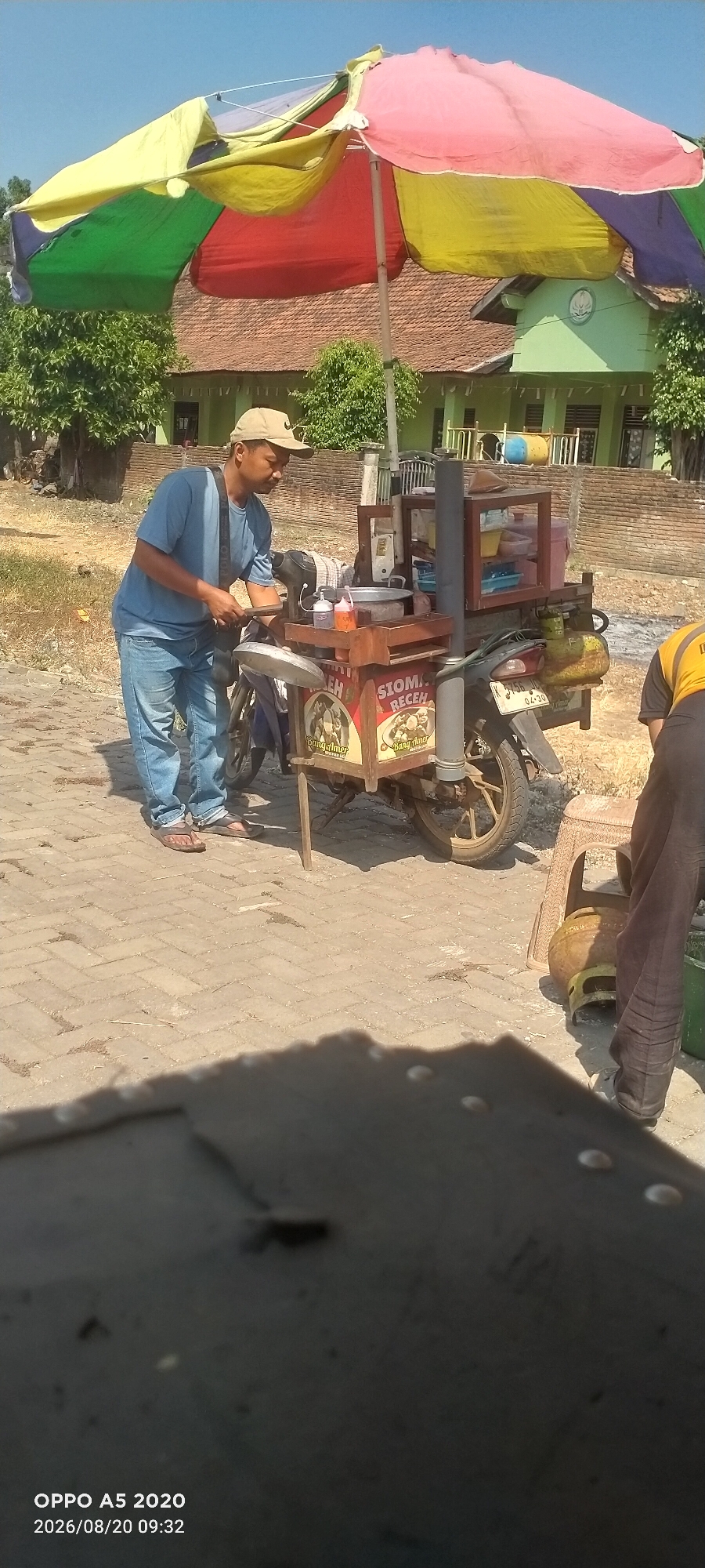 In Sowan Kidul, Indonesia, a street vendor is seen preparing food at a mobile stall attached to a motorcycle. The vendor, wearing a light blue t-shirt, jeans, and a beige baseball cap, is focused on their work. The stall itself is wooden and mounted on the back of a motorcycle, featuring several containers for ingredients, bottles of condiments, and a silver pot. A colorful umbrella, segmented with red, yellow, green, and purple, provides shade over the stall. In the background, a residential building with a tiled roof and green walls is visible, along with trees and a brick wall. Another person, partially visible and wearing dark pants and a yellow shirt, is near a propane tank and a plastic chair. The scene is set on a paved area with a herringbone brick pattern. The foreground is dominated by a dark, textured surface that partially obscures the view. The image captures a moment of daily commerce in this Indonesian town.