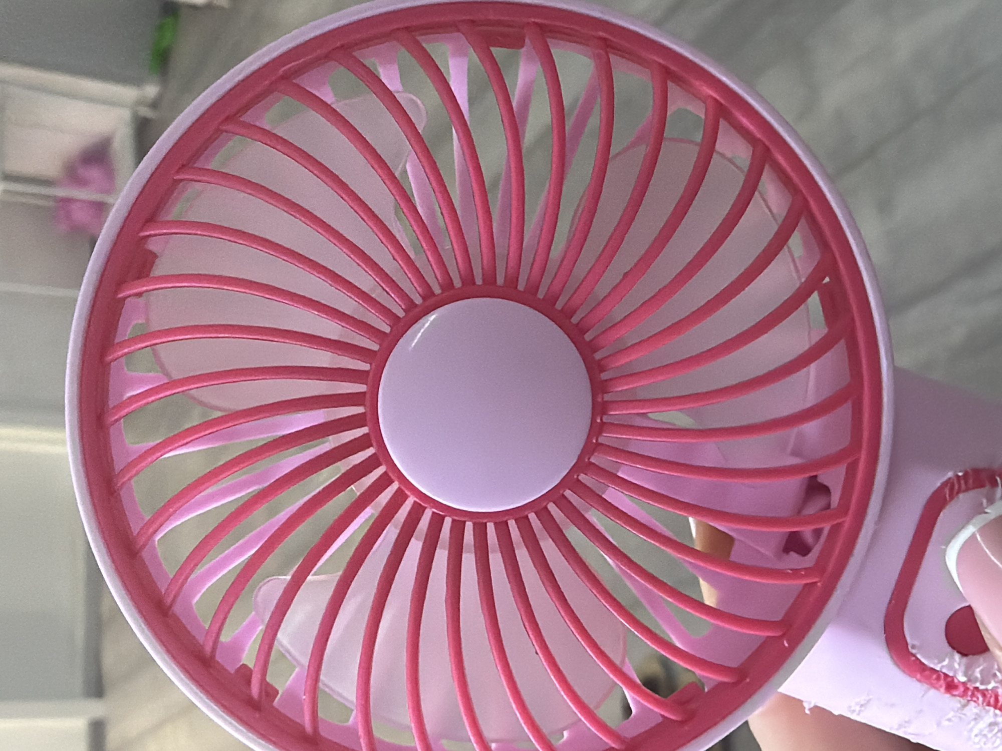 A close-up, eye-level shot shows a pink, portable fan with a central white button. The fan has a circular grille with thin, bright pink vertical bars radiating from the center. Behind the grille are translucent pink fan blades. The fan is held by a person's hand, with a pink fingernail visible on the left side of the frame. The background is blurred, suggesting an indoor setting with a light-colored floor or surface. The image is a still photograph with no visible action or interaction occurring. The location is not determinable from the visual information provided.