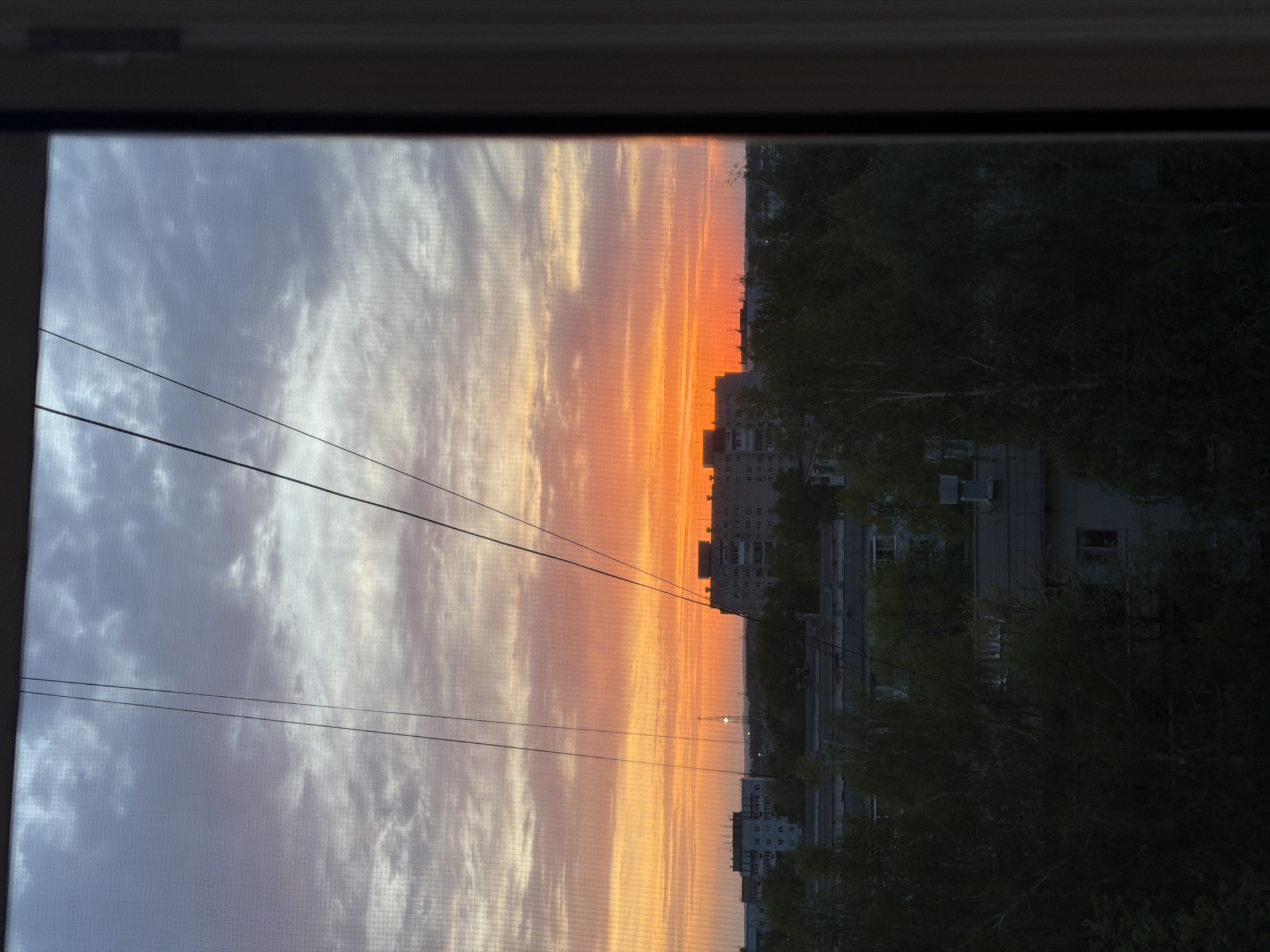 The image shows an urban scene viewed through a window in Chaikovsky, Russia, likely an apartment window due to the mesh screen visible. The sky transitions from a cloud-filled, pale light on the left to a vibrant orange on the right, indicative of a sunset. In the distance, silhouettes of buildings and trees mark the cityscape. Power lines are strung across the window's view of the sky.