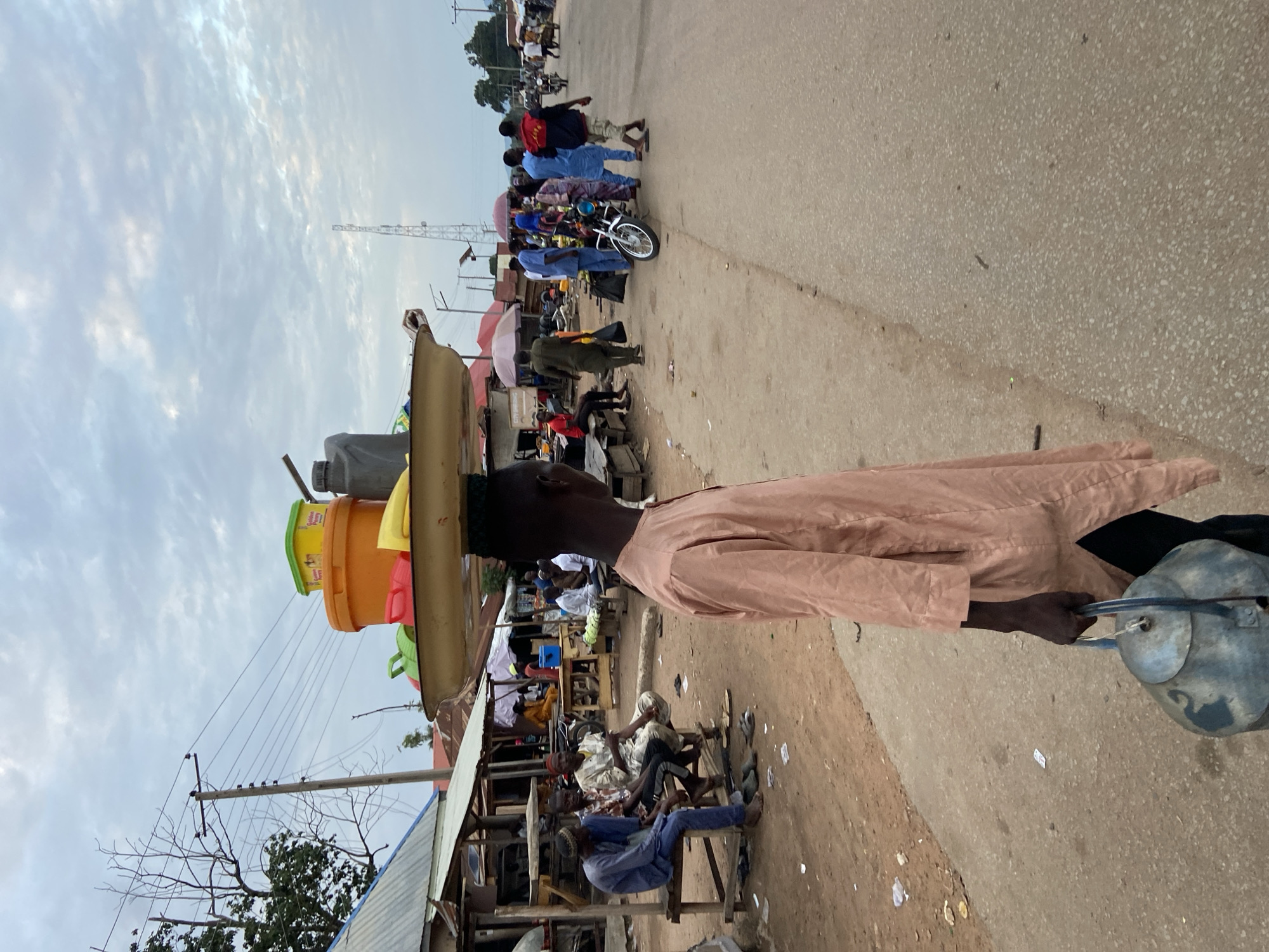 A man is carrying a stack of plastic containers on his head, walking on a road in the city of Mararaba, Nigeria. He is wearing a light brown, long-sleeved shirt. To his right, there are various people, including a group of men sitting on wooden benches and chairs in front of market stalls. In the background, a group of people are gathered around a motorcycle. The sky is cloudy and there is a tall communication tower visible. The road is paved but also has dirt and debris along the edge.
