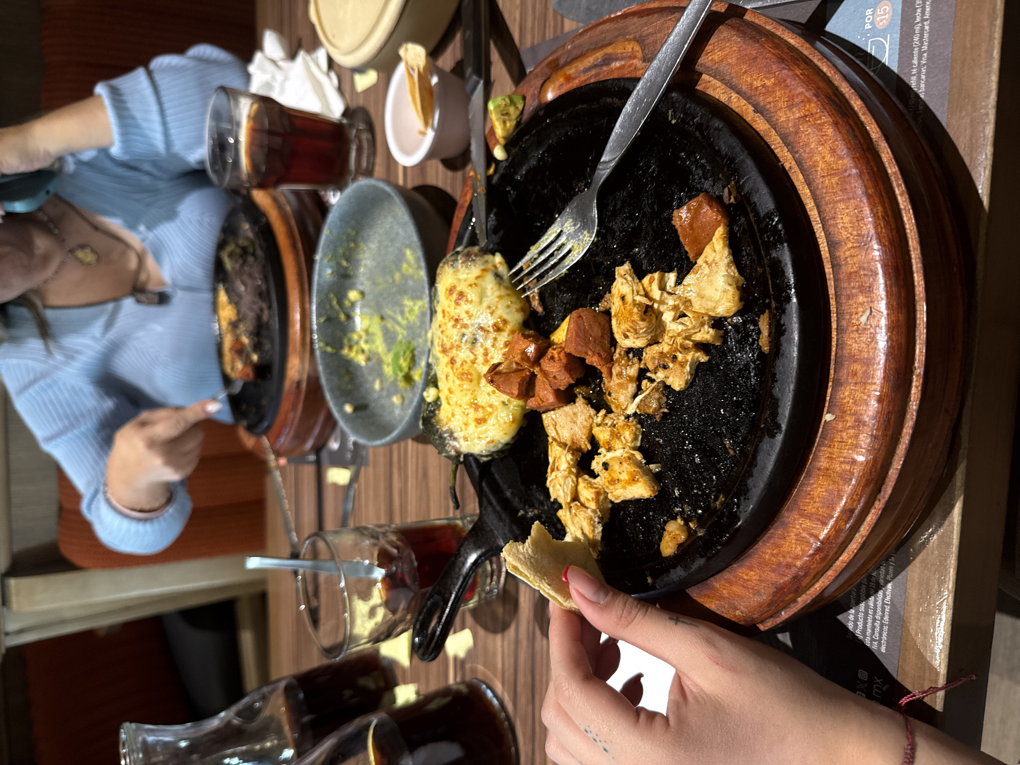 The image displays an indoor dining scene at a restaurant in Puebla, Mexico. The foreground features a person's left hand, adorned with red nail polish on the thumb and index finger, a small cross tattoo on the wrist, and a red string bracelet. This hand holds a small, irregularly shaped piece of light-colored flatbread.
The primary subject is a black cast-iron sizzle platter, resting on a wooden base, positioned centrally. The platter contains several pieces of cooked chicken, reddish-brown chunks identified as chorizo, and a large green chili covered in melted white cheese, likely a chile relleno. A silver fork with its tines facing left is partially submerged in the food on the platter.
To the immediate left of the platter, a gray bowl holds green remnants consistent with guacamole. To the right, a small white bowl contains a few yellowish fragments, possibly tortilla chips. Two clear glass carafes and two clear glasses, all containing a dark brown liquid, are arranged on the wooden table surface. One glass has a long spoon inserted.
In the mid-background, a second individual is visible from the chest up, wearing a light blue ribbed long-sleeved top with a v-neck and a necklace with a pendant. This person is actively cutting food on another black cast-iron platter using a fork and knife.
The environment consists of a wooden tabletop, a visible orange upholstered chair in the background, and ambient lighting. A placemat or menu partially visible in the bottom left and right corners includes text referring to "Puebla" and a price of "POR $15", supporting the identified location and local currency context.