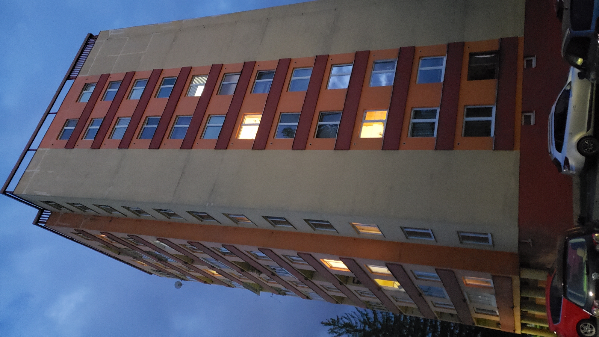 The image shows a residential high-rise building in Prešov, Slovakia. The building is tall and narrow, with multiple floors of apartments. The exterior is painted in a combination of beige and orange, with dark red horizontal accents. Many of the windows are illuminated, suggesting that it is evening or night and residents are home.
The building has a dark-colored railing around the rooftop. There are some satellite dishes visible on the side of the building. A dark green conifer tree is partially visible on the left side of the image.
At the base of the building, there are several parked cars. A red car is parked on the far left. To its right is a dark-colored car, possibly a hatchback. Further to the right is a silver station wagon. The ground in front of the building appears to be a paved area or sidewalk. The sky is a deep blue, indicating dusk or twilight.
The building appears to be an apartment block, common in urban areas like Prešov. The presence of parked cars and illuminated windows suggests it is a populated residential area. The architectural style is typical of Soviet-era or post-Soviet era apartment buildings. The overall impression is of a typical urban residential scene at the end of the day.