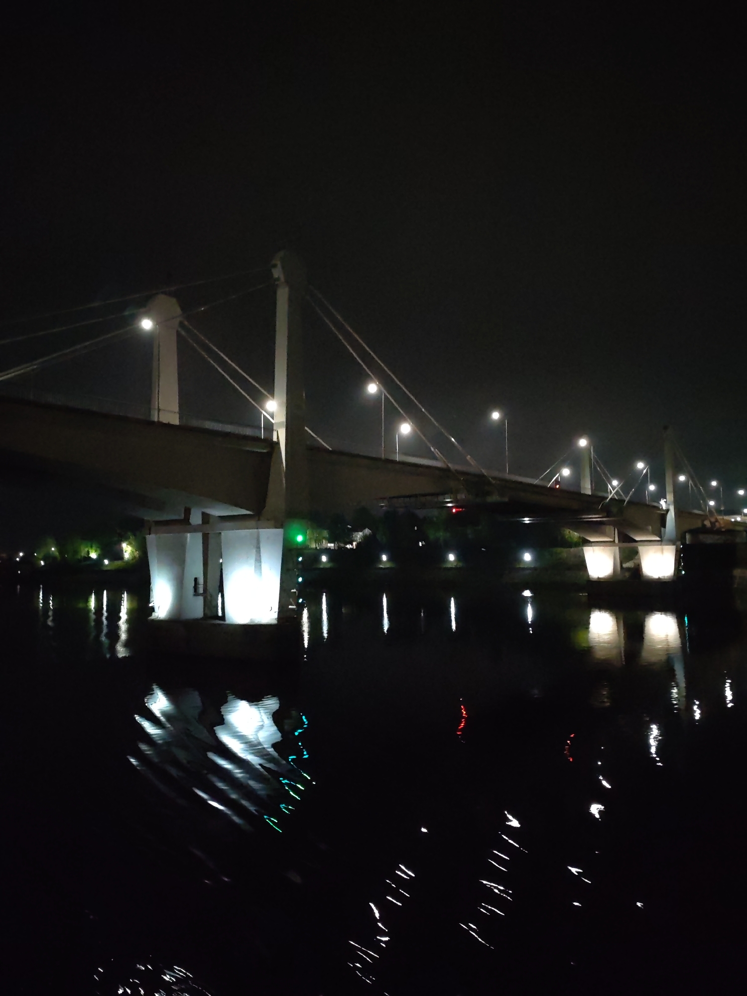At night, a bridge in Kimry, Russia is illuminated with multiple lights, casting reflections on the dark water below. The bridge has a cable-stayed design with tall, white pillars. Lights line the bridge, and there are trees and vegetation on the nearby land. The water surface is disturbed, creating fragmented reflections of the lights.