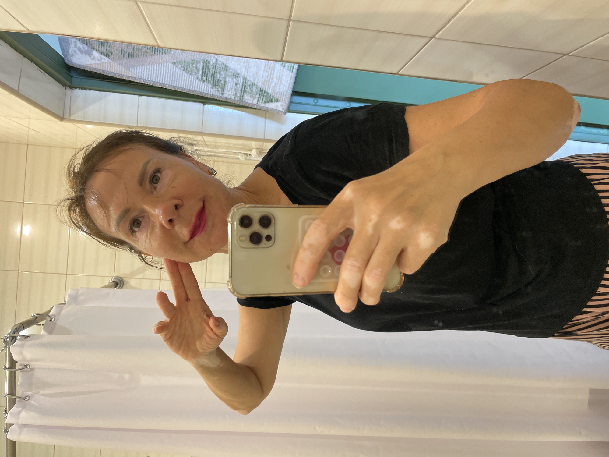 A woman is taking a selfie in a bathroom. She is wearing a black t-shirt and striped pants. Her hand is raised to her face, and she is looking at the camera. The background shows a tiled wall, a shower curtain, and a door. The scene is lit with artificial light. There is no indication of the city of Sochi, Russia, in the image.