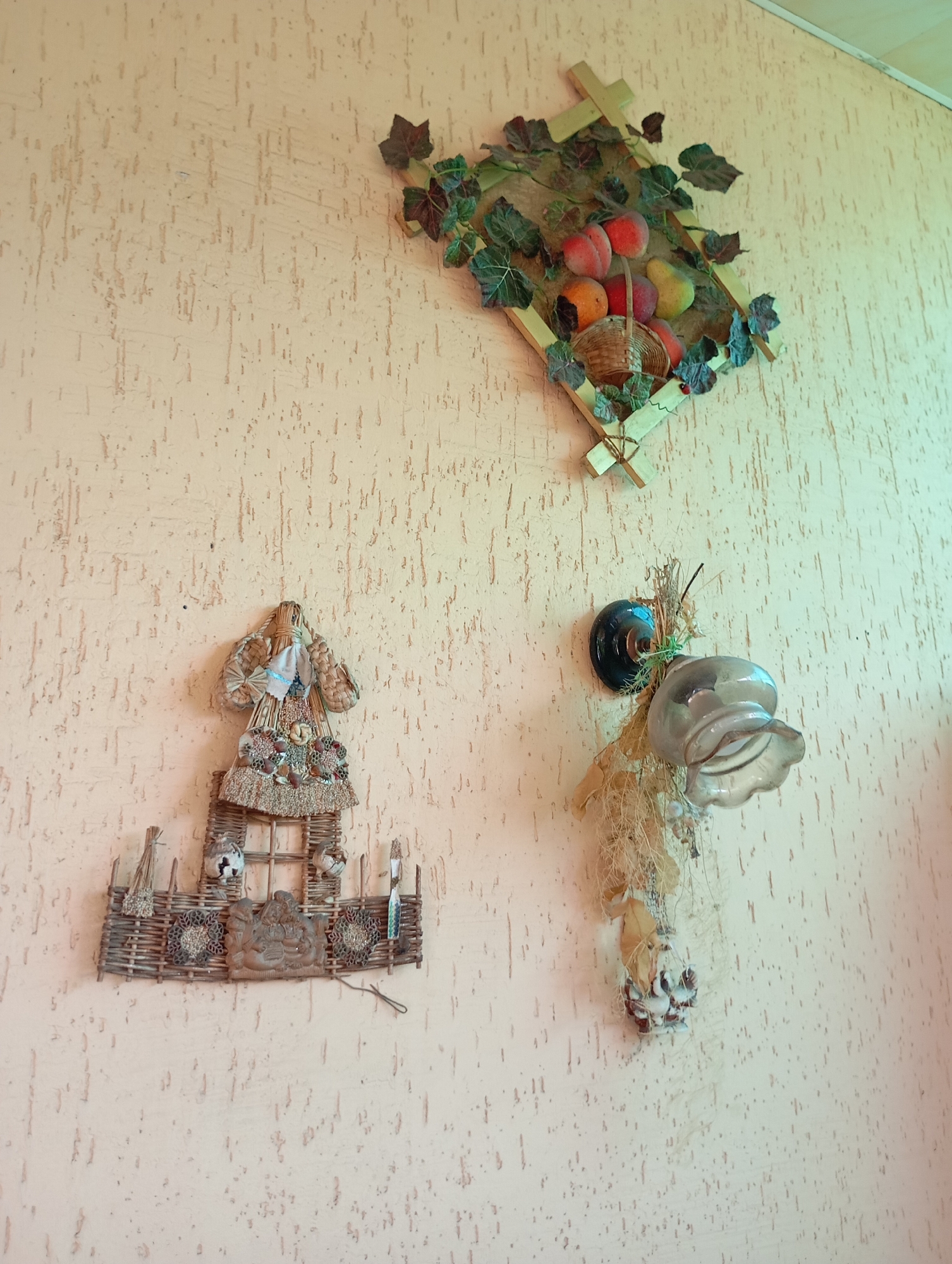 The image shows a textured beige wall with three decorative items. At the top, a diamond-shaped wooden frame is adorned with artificial green leaves and a cluster of faux fruit, including peaches and pears, with a small basket. Below and to the left, a straw doll, possibly a folk art representation, is depicted wearing a straw hat and dress decorated with shells and other natural elements. It is mounted on a woven straw structure resembling a fence with small bird figures and decorative elements. To the right of the straw doll, a glass object, perhaps a sconce or decorative light fixture, is hanging. It is made of smoky glass and is adorned with dried grasses and natural materials. The overall scene suggests a rustic or folk-inspired interior decoration, likely in a residential setting in Horishni Plavni, Ukraine.