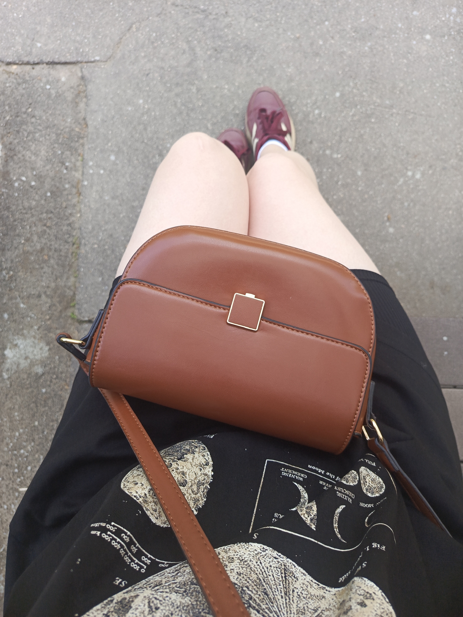 The image depicts a person sitting outdoors, likely on a bench or ledge, as suggested by the gray concrete visible in the background. The individual's legs are visible, with maroon sneakers on their feet. A brown handbag with a gold square clasp rests on their lap. The person is wearing a black shirt with a white lunar phase design and text, part of which reads "WAXING CRESCENT" and "WANING CRESCENT." The location is unknown and not verifiable.