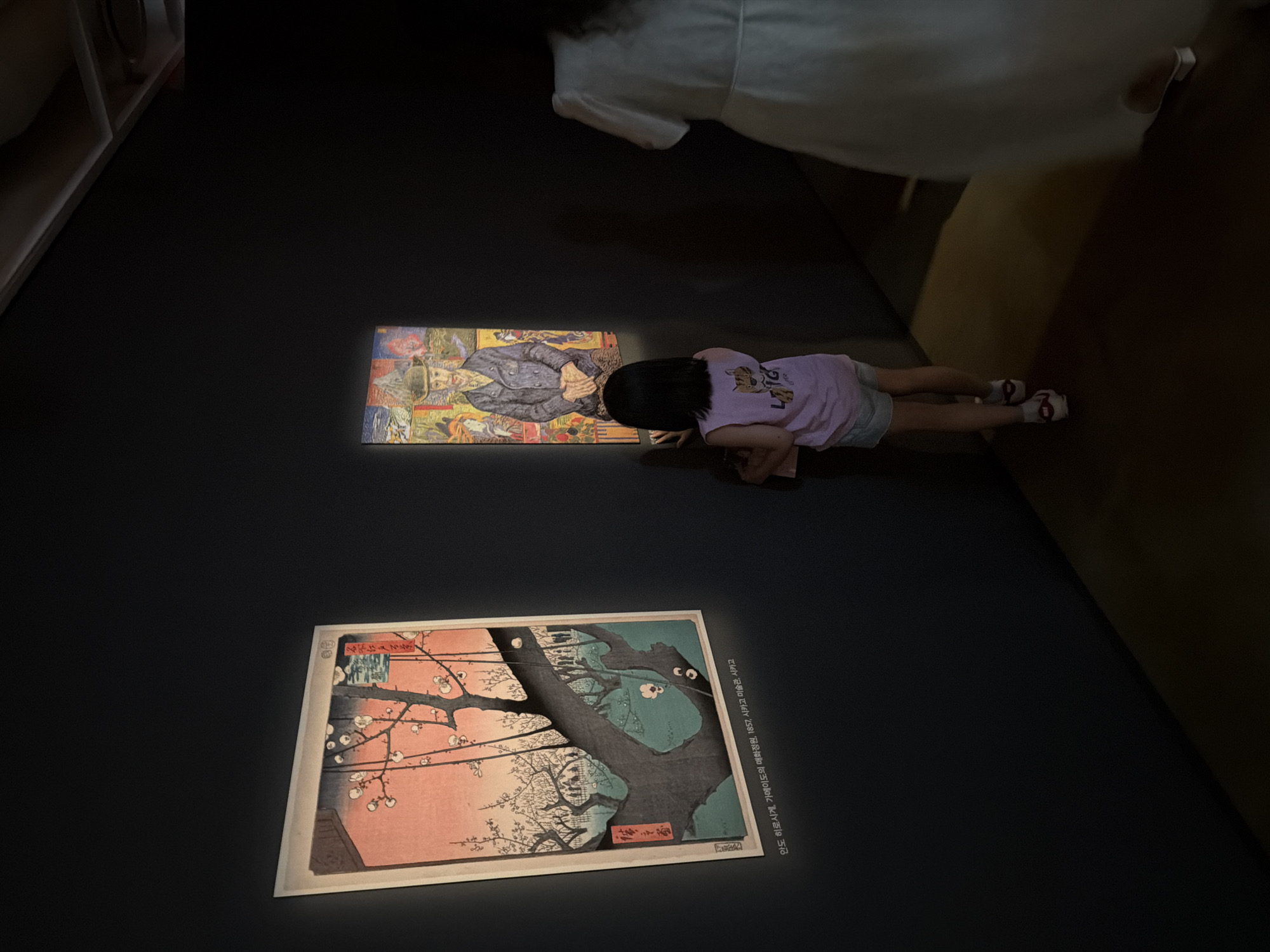 The image depicts an indoor scene, likely an art gallery or museum, with dark walls and artificial lighting. In the foreground, a young girl with dark hair, wearing a pink t-shirt and denim shorts, is looking at a framed artwork on the left wall. The artwork appears to be a traditional Japanese woodblock print depicting a tree with pink blossoms and a vibrant pink and blue sky. Below this artwork, a caption in Korean is visible. To the right of the girl, another framed artwork is displayed, featuring a portrait of a man in a hat and suit, surrounded by a colorful, fragmented background. Further to the right and slightly blurred, a person in a long white dress is walking away from the viewer. The floor is a dark, reflective surface. The overall ambiance is quiet and focused on the art. Based on the Korean text accompanying the artwork on the left, the location is likely the Seoul Museum of Art, given the mention of "시카고 미술관" which translates to "Chicago Museum of Art," indicating an exhibition that may have originated or been influenced by works from that institution, and the context of a museum in Seoul.