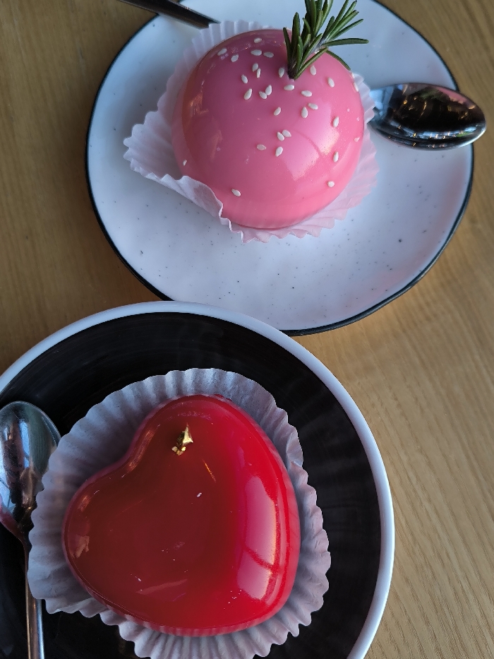 The image displays two individual desserts presented on a light wooden table, consistent with a cafe or restaurant setting in Vinnytsia, Ukraine. The upper dessert is a spherical, glossy pink mousse cake, topped with white sesame seeds and a green rosemary sprig, resting in a white fluted paper liner on a speckled white ceramic plate with a black rim. A silver spoon lies to the right of this plate. The lower dessert is a heart-shaped, glossy red mousse cake, garnished with a small gold leaf, also resting in a white fluted paper liner on a black ceramic plate with a white rim. A second silver spoon is positioned to the left of the lower plate. No individuals are visible in the scene. The items are illuminated by overhead or ambient light, producing reflections on the desserts and cutlery.