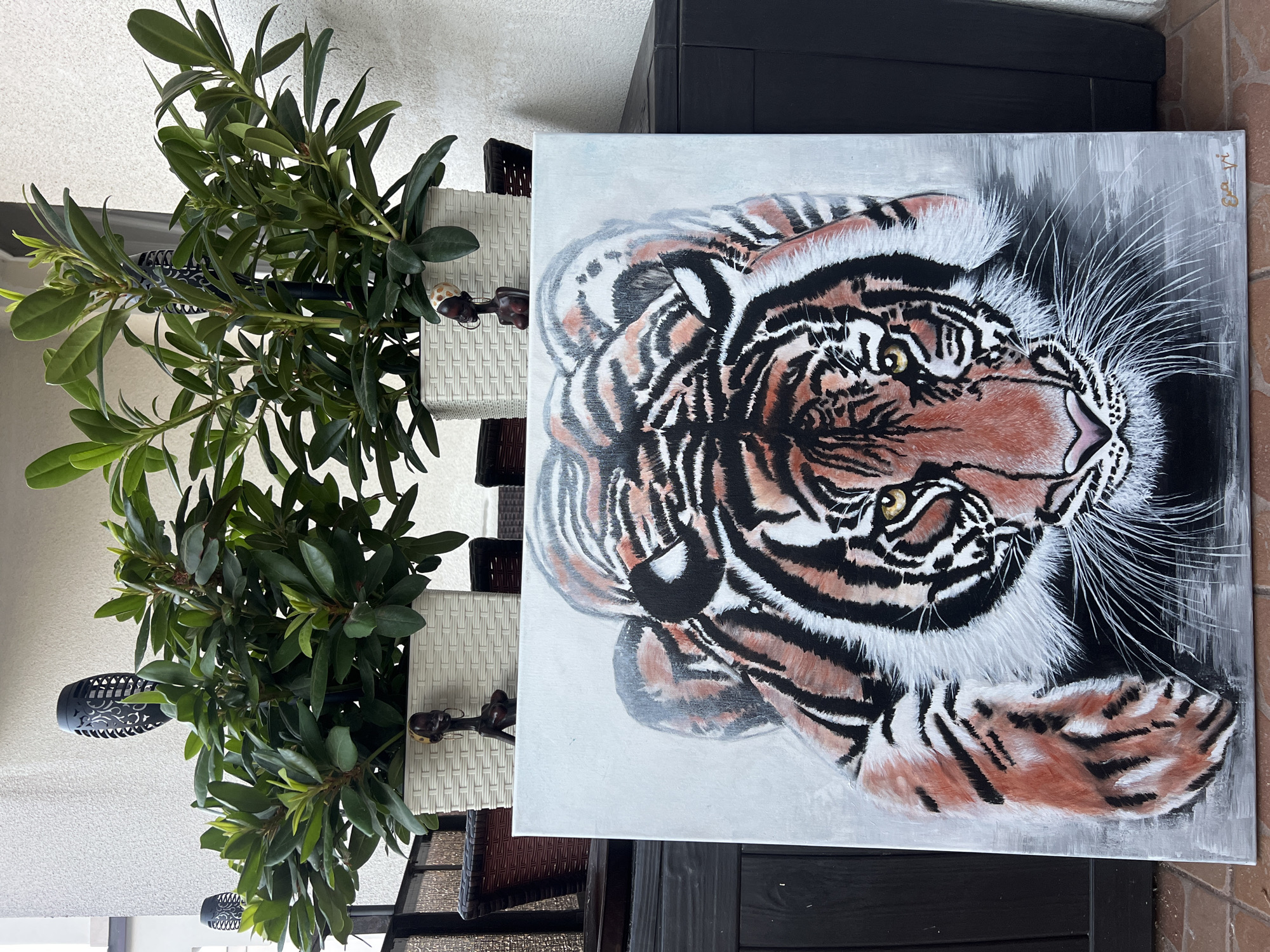 An oil painting of a tiger in a stalking pose is displayed outdoors on a dark wooden surface. The painting is a close-up of the tiger's head and front paws, with its eyes focused forward. The tiger's fur is depicted with orange, black, and white stripes, and its whiskers are white and sparse. The background of the painting is a light gray.
Behind the painting, two white planters with lush green foliage are visible. Each planter is adorned with a small, dark wooden figurine of a woman with her arms crossed and wearing a yellow headscarf. The planters are placed on a dark surface, possibly a railing or a piece of furniture. In the background, a textured, light-colored wall is visible, and a black solar-powered lamp with a cut-out design is placed behind the foliage in the left planter. The ground beneath the painting appears to be tiled with reddish-brown bricks.
The scene is set outdoors, likely on a patio or balcony. The presence of planters and outdoor furniture suggests a residential setting. While the precise location within Kleosin, Poland, is not explicitly stated, the overall ambiance points to a domestic outdoor space.