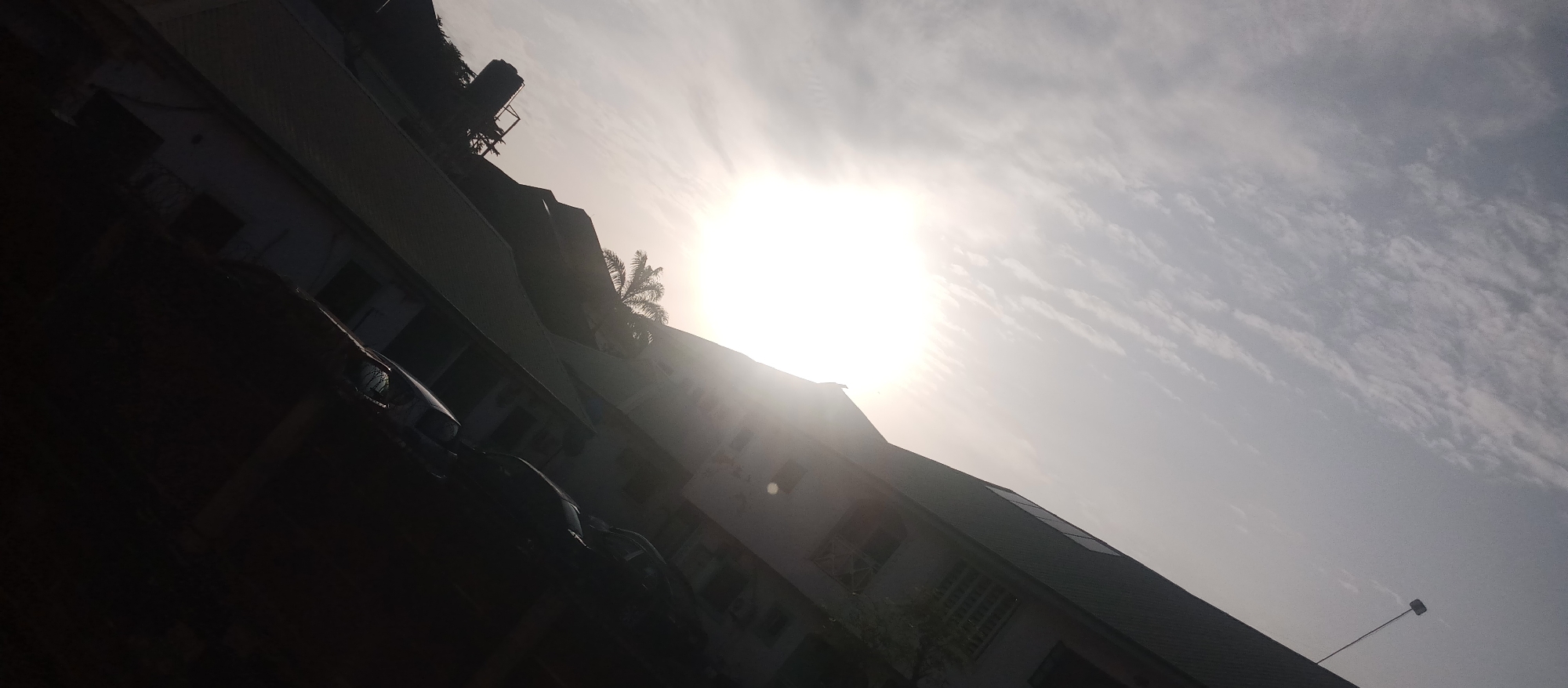 An eye-level, slightly angled, exterior shot captures a sunlit sky and a silhouetted building with cars parked in front. The sun is prominently featured, emitting bright rays that cast shadows on the scene. A water tank is visible atop the building. A lone palm tree stands to the right of the frame. The image is dominated by the bright sky and the dark silhouette of the building and cars.