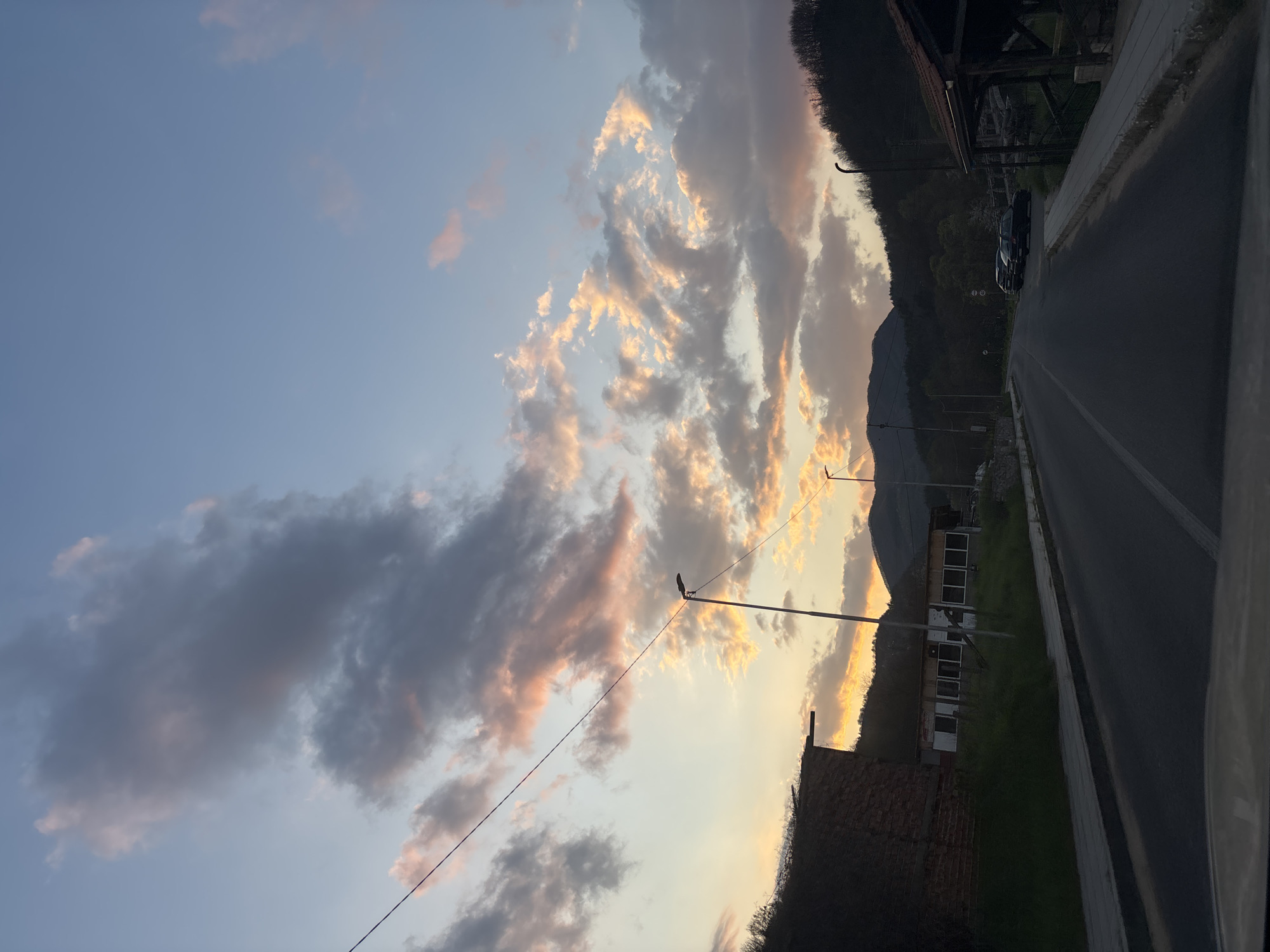 The image shows a street scene in Zlatograd, Bulgaria, at dusk. The sky is filled with clouds, some tinged with orange and pink from the setting sun. A road runs along the right side of the frame, with a sidewalk to its left. A building with white-framed windows is visible along the road. A utility pole stands near the building, and a power line stretches across the sky. Further down the road, cars are parked near a covered structure. The mountains are visible in the background.