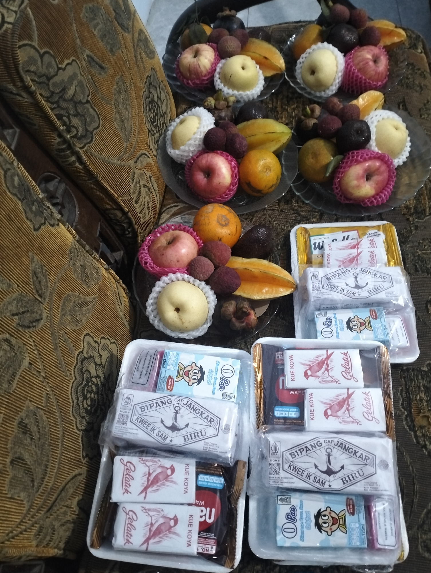 The image displays an assortment of fruits and packaged snacks arranged on a patterned couch. The fruits, including apples, lychees, star fruit, oranges, mangosteens, and pears, are presented in clear plastic bowls. Below the fruit, multiple white rectangular trays hold various packaged snacks. These snacks include items with wrappers featuring branding such as "BIPANG CAP JANGKAR," "KWE IK SAM BIRU," "Gelatik," "KUE KOYA," and small juice boxes with cartoon characters. The overall arrangement suggests a gift or a prepared assortment for sharing. The couch's brown and beige floral pattern indicates an indoor setting. There are no people visible in the image. The location is specified as Pleret, Indonesia.