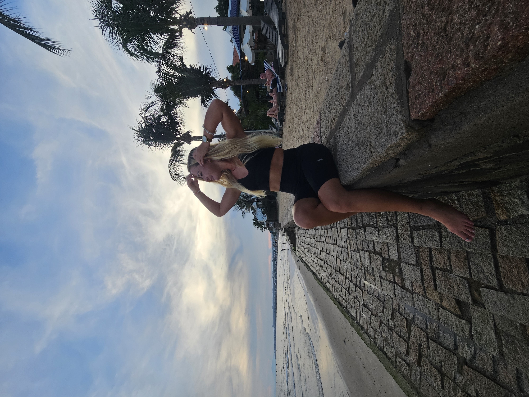 A blonde woman in athletic wear sits on a stone ledge at the edge of a beach in Phu Thuy Ward, Vietnam. The woman is facing left, with her hands raised to her hair. She is wearing a black sports bra and black biker shorts. To her right, on the sand, are several lounge chairs. Palm trees and a building are visible in the background. The sky is cloudy with hints of orange and pink, suggesting it is either sunrise or sunset. The foreground shows a textured stone walkway leading to the beach. The ocean is to the left of the image.