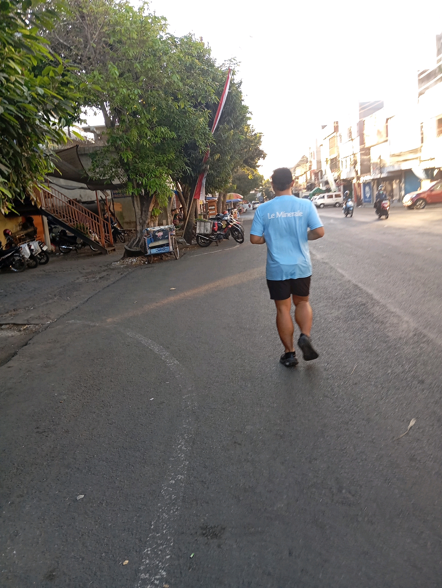 A man with muscular calves runs on a paved road in what appears to be a city in Malang, Indonesia. He is wearing a light blue t-shirt with "Le Minerale" printed on the back and black shorts. The road is lined with trees and buildings on the right, and parked motorcycles and a small building with a metal staircase on the left. The sun is low in the sky, casting long shadows and creating a bright, hazy atmosphere. A few other motorcycles and a car can be seen in the background.