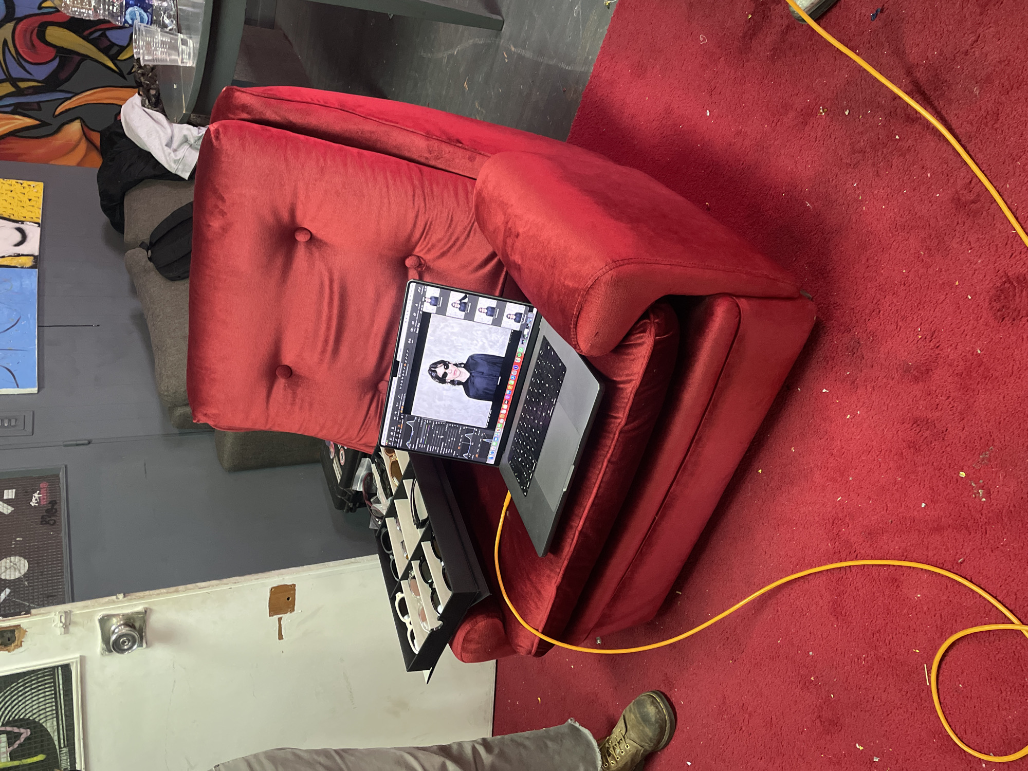 A laptop sits atop a red velvet armchair on a red carpet. An open case of eyewear sits on the arm of the chair. An orange cord runs from the laptop to the floor. A person wearing khaki pants and boots stands partially in frame. The room is cluttered. Artwork is visible on the wall.
There is not enough information to determine if the photo was taken in Buenos Aires, Argentina.