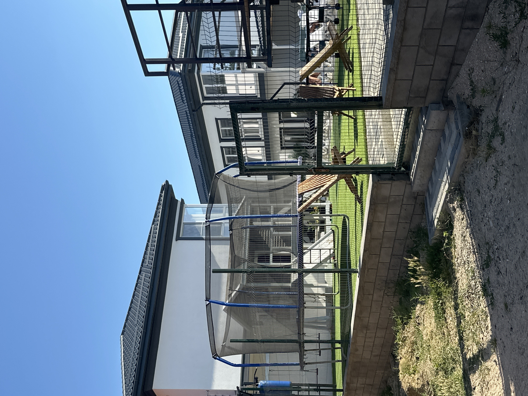 In Golubitskaya, Russia, a residential backyard is depicted on a clear, sunny day. The scene features two modern, multi-story houses with dark tiled roofs and white exteriors, separated by a manicured green lawn. In the foreground, a tall wire fence encloses the property, with a gate visible. Inside the fenced area, a large trampoline with blue poles and a safety net stands prominently. To the right of the trampoline, wooden lounge chairs and playground equipment, including slides, are scattered across the lawn. The ground outside the fence is a mixture of dirt, sand, and sparse vegetation, with a low brick wall bordering the lawn. The overall impression is one of a well-maintained, recreational outdoor space associated with a residential dwelling.