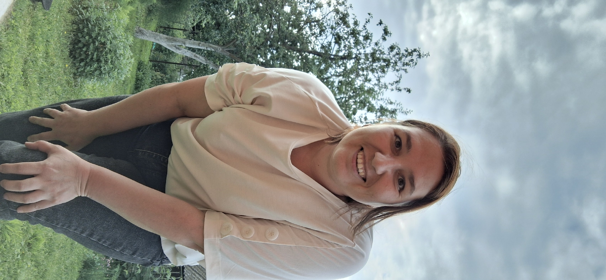 A medium shot portrait features a woman with light brown hair, a fair complexion, and a broad smile. She is wearing a light-colored t-shirt with decorative buttons on the sleeves and dark denim jeans. The woman is outdoors, with her hands resting on her knees, in a slightly bent posture. Behind her, a tree with green foliage and a cloudy sky are visible. The background suggests a residential or park-like setting, consistent with the environment found in Chaikovsky, Russia.