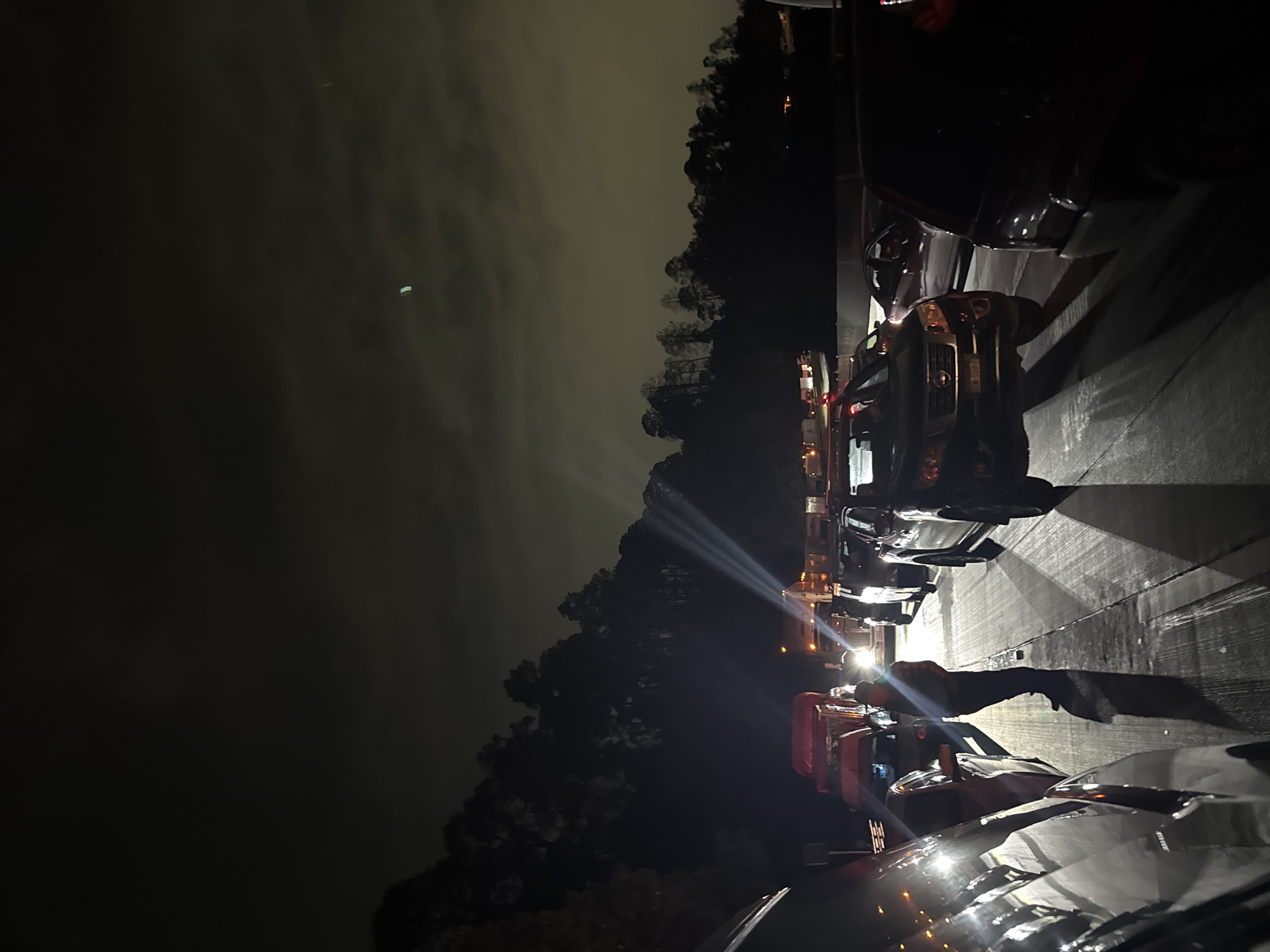 The image presents a nighttime scene in Llano Grande, Mexico, featuring several vehicles parked or arranged along a dark, paved surface, likely a road or parking lot. The overall lighting is low, with artificial light sources dominating the visibility.
On the right side of the frame, multiple cars and what appear to be large trucks are lined up. A prominent red truck is visible with its lights on. The vehicles' headlights and auxiliary lights are brightly illuminated, casting strong beams and reflections that stretch across the ground, creating distinct patterns of light and shadow.
A clear silhouette of a person, likely an adult, stands near the vehicles on the right. This individual casts a long, exaggerated shadow onto the ground, indicative of a bright light source positioned behind them. Other, less distinct figures might also be present further back among the vehicles.
The background is dominated by a dense line of trees and foliage, appearing as dark silhouettes against the sky. The night sky itself is overcast, with clouds showing a lighter grey tone, suggesting they are reflecting ambient light or diffusing light from above. This creates a soft glow in the upper left portion of the image.
The scene is dark and atmospheric, characterized by the interplay of artificial lights against the natural darkness of the night. No discernible text is visible on any objects or in the environment.
