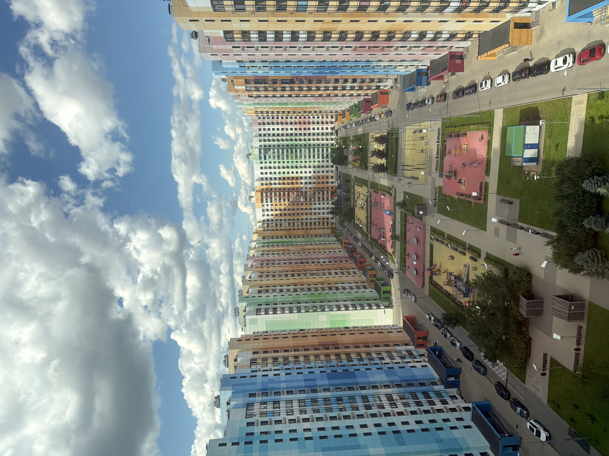 The image shows a high-angle view of a modern residential complex with buildings of varying colors, including blue, green, pink, orange, and yellow. The buildings are tall and narrow, with numerous windows and balconies. In between the buildings are courtyards with playgrounds, green spaces, and paved walkways. Several cars are parked along the street that runs through the complex. The sky is partly cloudy.