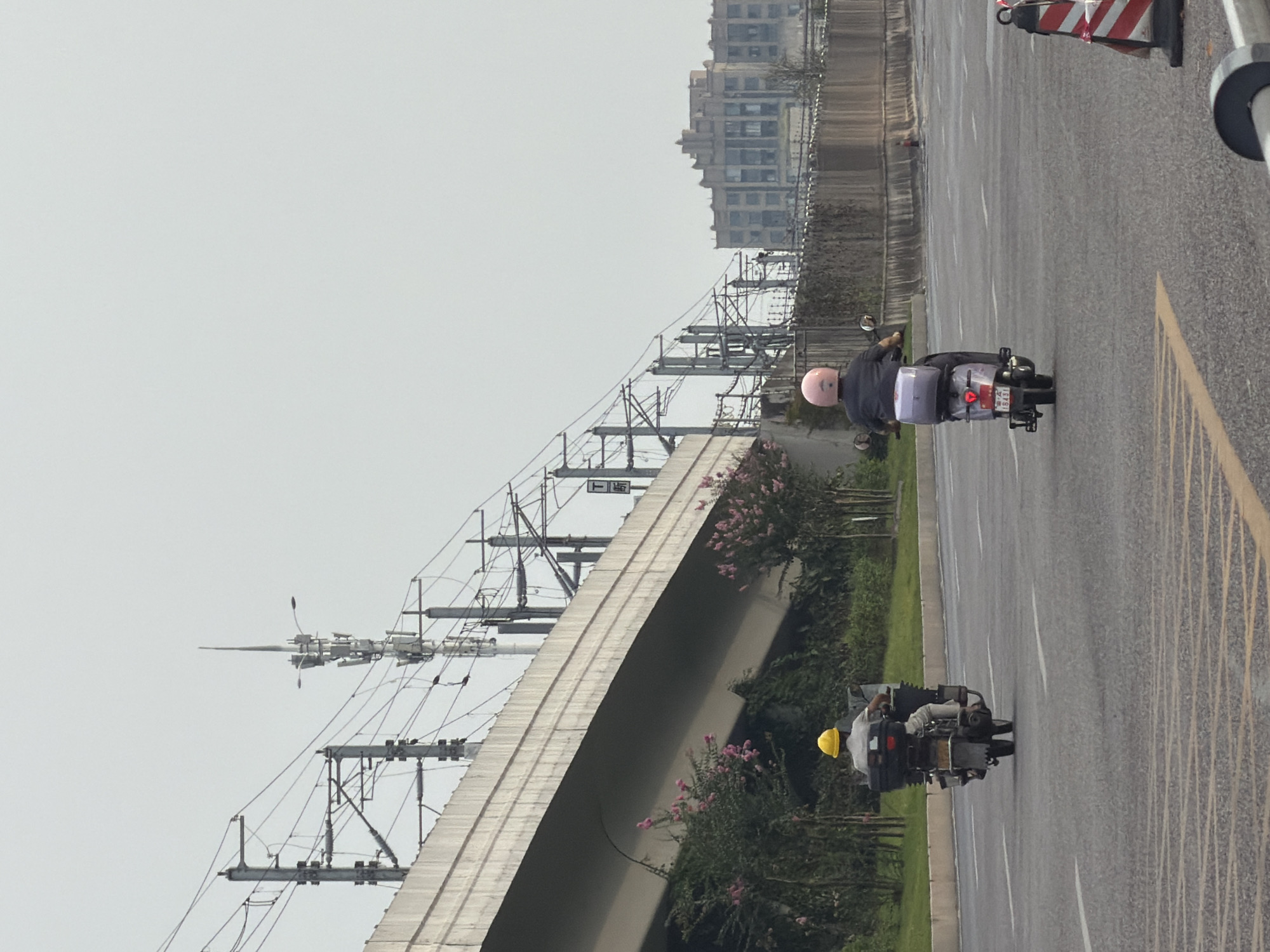Two individuals are riding motorbikes on a road in Jiangjin, China. The rider in the foreground is wearing a pink helmet and a dark jacket, with a light-colored storage box on the back of their motorbike. The rider further in the distance is wearing a yellow hard hat and a light-colored shirt, also with a dark storage box on their motorbike. The road is paved with asphalt and marked with white lane lines. To the left of the road, a raised concrete structure, likely an overpass or elevated road, is visible. Overhead, power lines and communication towers are present. Behind the concrete structure, lush greenery and flowering bushes can be seen. In the background, a large, multi-story building with a distinctive architectural style stands against a hazy sky. The overall scene suggests an urban or semi-urban environment.