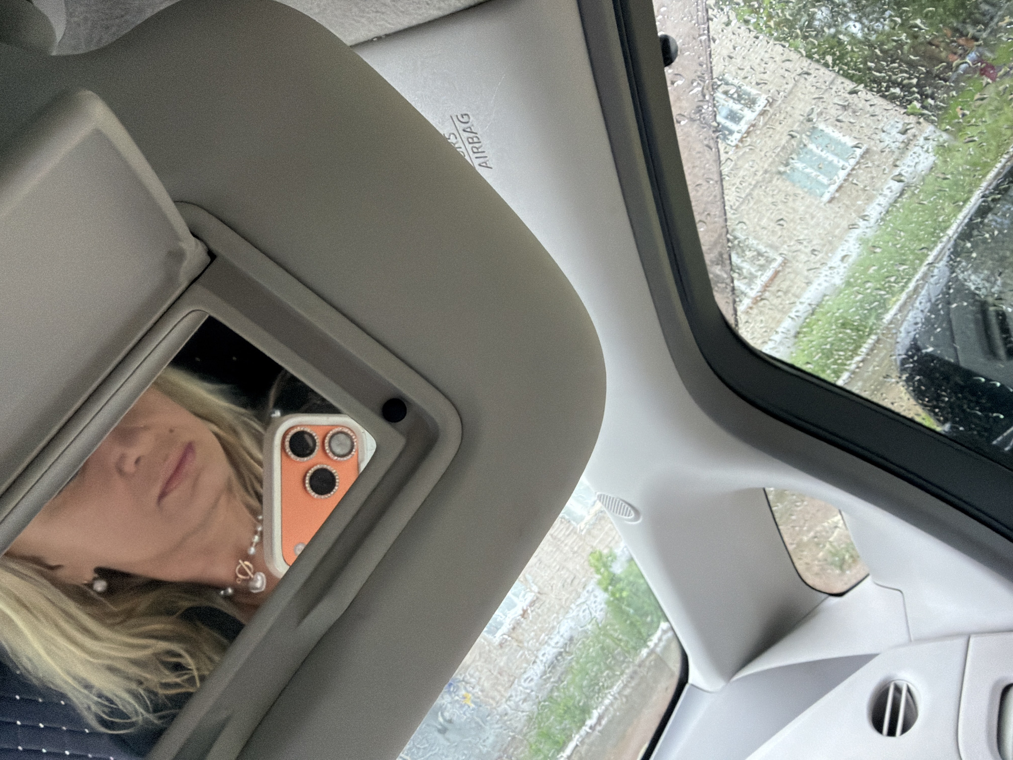 The image shows the interior of a car, likely in Chaikovsky, Russia, as indicated by the reflections in the car window of buildings typical of Russian urban areas. Rain droplets cover the car window, partially obscuring the view of the buildings and surrounding greenery outside.
A sun visor with a mirror is positioned on the driver's side of the car. The mirror reflects a partial view of a woman with blonde hair, pearl earrings, and a pearl necklace. She is holding a cell phone with an orange case, decorated with circular jewels, that is also reflected in the mirror.
The car's interior is light grey. A section of the seat, visible below the visor, appears to be upholstered with a dark-colored fabric with a dotted pattern. An "AIRBAG" warning label is printed on the sun visor. An air vent is visible near the edge of the car's windshield. The scene is static. There are no apparent actions or interactions occurring.