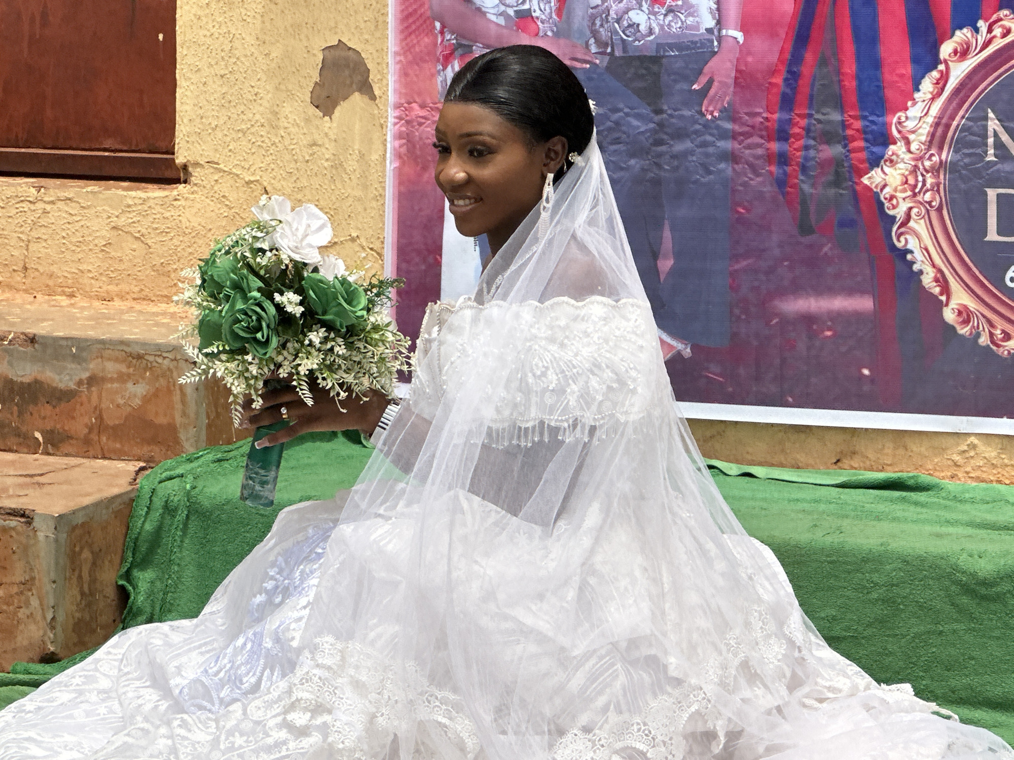 A bride in Unknown, Unknown, sits holding a bouquet of green and white flowers on a green-covered surface, with a backdrop of peeling yellow paint and a blurred advertisement featuring red and blue stripes and ornate framing. The bride, wearing a white wedding dress and veil, gazes to the side with a slight smile, and her appearance is completed by hanging earrings and dark, neatly styled hair.