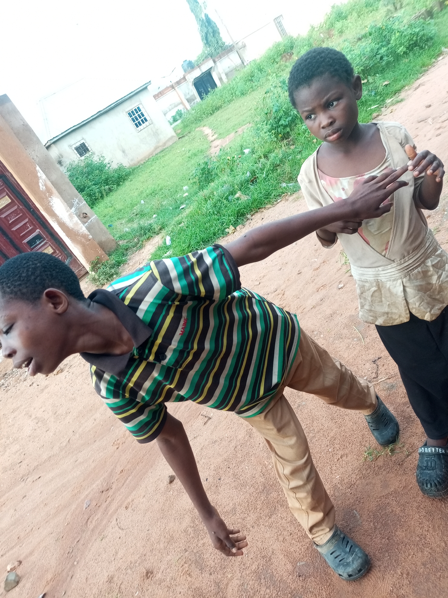Two young children, a boy and a girl, are standing on a dirt path in an outdoor environment. The boy, in the foreground, is wearing a striped shirt with green, yellow, black, and white bands, and khaki pants. He is leaning to his left, with his eyes closed and his mouth open, in what appears to be a playful or dramatic pose. His right arm is extended forward. The girl, in the background, is wearing a light-colored, possibly beige, short-sleeved top with some smudges on it, and black pants. She is looking to her left with a serious expression and has her right hand extended, touching the boy's outstretched arm. The background shows a building with a light-colored facade and a metal roof, as well as some green vegetation. The overall setting suggests a rural or semi-urban area in Tempure, Nigeria.