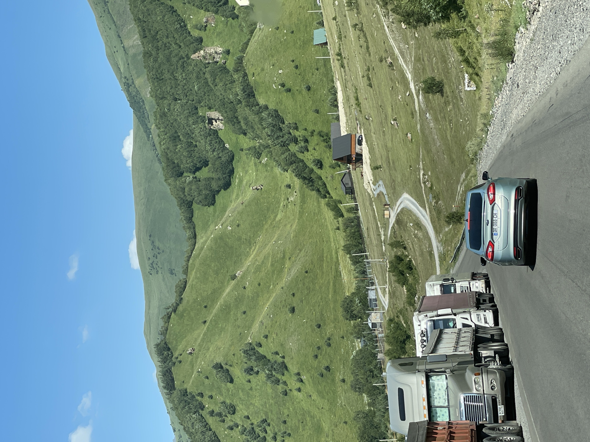 The image depicts a road in the mountainous region near Sioni, Georgia, on a clear day. A line of trucks is parked on the right side of the road, and a light blue car is in the foreground, driving away from the viewer. The road winds through a lush green valley with rolling hills covered in grass and scattered trees. In the background, several houses and buildings are visible, nestled into the landscape. The sky is bright blue with a few scattered white clouds.