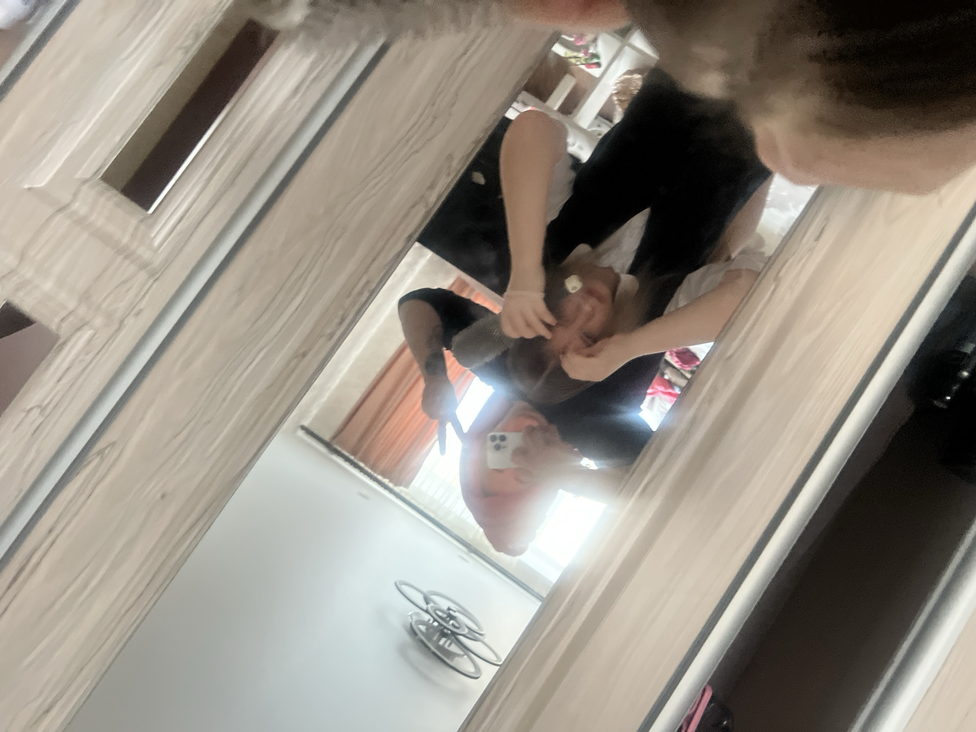 The image is a mirror selfie taken indoors, showing the reflection of a person from an overhead or angled perspective. The reflection appears upside down relative to the frame.
The **subject** is a young person, whose face is largely obscured by the white smartphone they are holding to capture the image. They are wearing dark clothing, possibly black pants and a dark top. One hand is visible, either holding the phone or making a 'peace' sign gesture.
The **activity** is taking a casual mirror selfie.
**Notable details** include the room's interior, which features light wood-grain patterned door frames or closet doors. In the reflected background, there are orange or brown curtains, a bright light source (likely a window) creating a significant glare, and shelving units with items on them. On the floor in the reflection, a modern, multi-ringed decorative object or light fixture is visible. The overall lighting suggests it is daytime. No discernible text is visible in the image. The setting appears to be a private indoor space, possibly a bedroom or dressing area, in Chaikovsky, Russia.