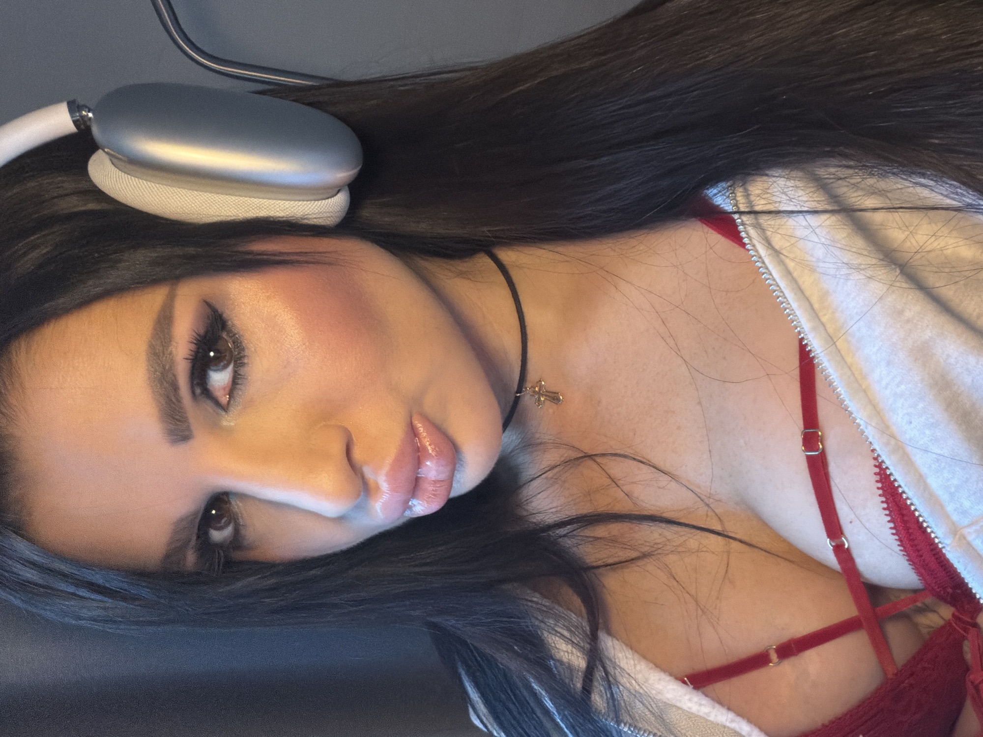 A close-up, angled view of a young woman with long dark hair wearing silver headphones and a choker with a small cross pendant. She is wearing a red bra under a white zippered hoodie. Her face is heavily made up with dramatic eye makeup and glossy lips. The background is dark and nondescript. The image does not show any details that would identify a location in Svkh Voronezhskiy, Russia, or any other specific place.