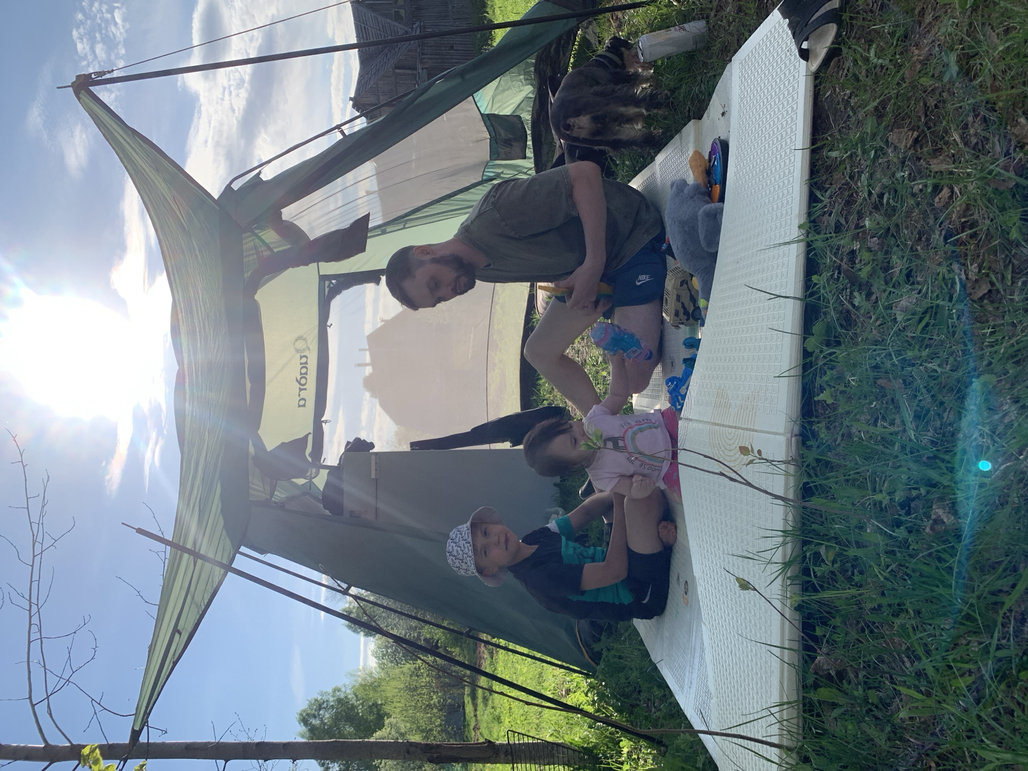 The scene depicts a family outdoors. A fair-skinned man with a beard is holding a blue water gun. Two young children, a boy wearing a patterned bucket hat and a girl in a pink shirt, are seated. They are on a light-colored, rectangular, foam-like surface. Above them is a tent-like structure. The location appears to be rural with grass. It is Zasechnyi, Russia.
